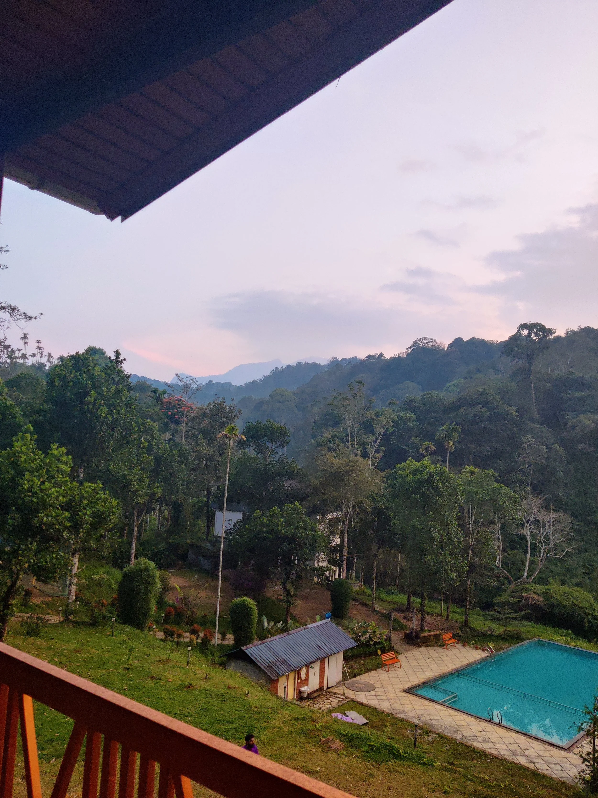 View of a mountain landscape with trees and hills, a swimming pool, a small building, and a patio area with benches, taken from a balcony.