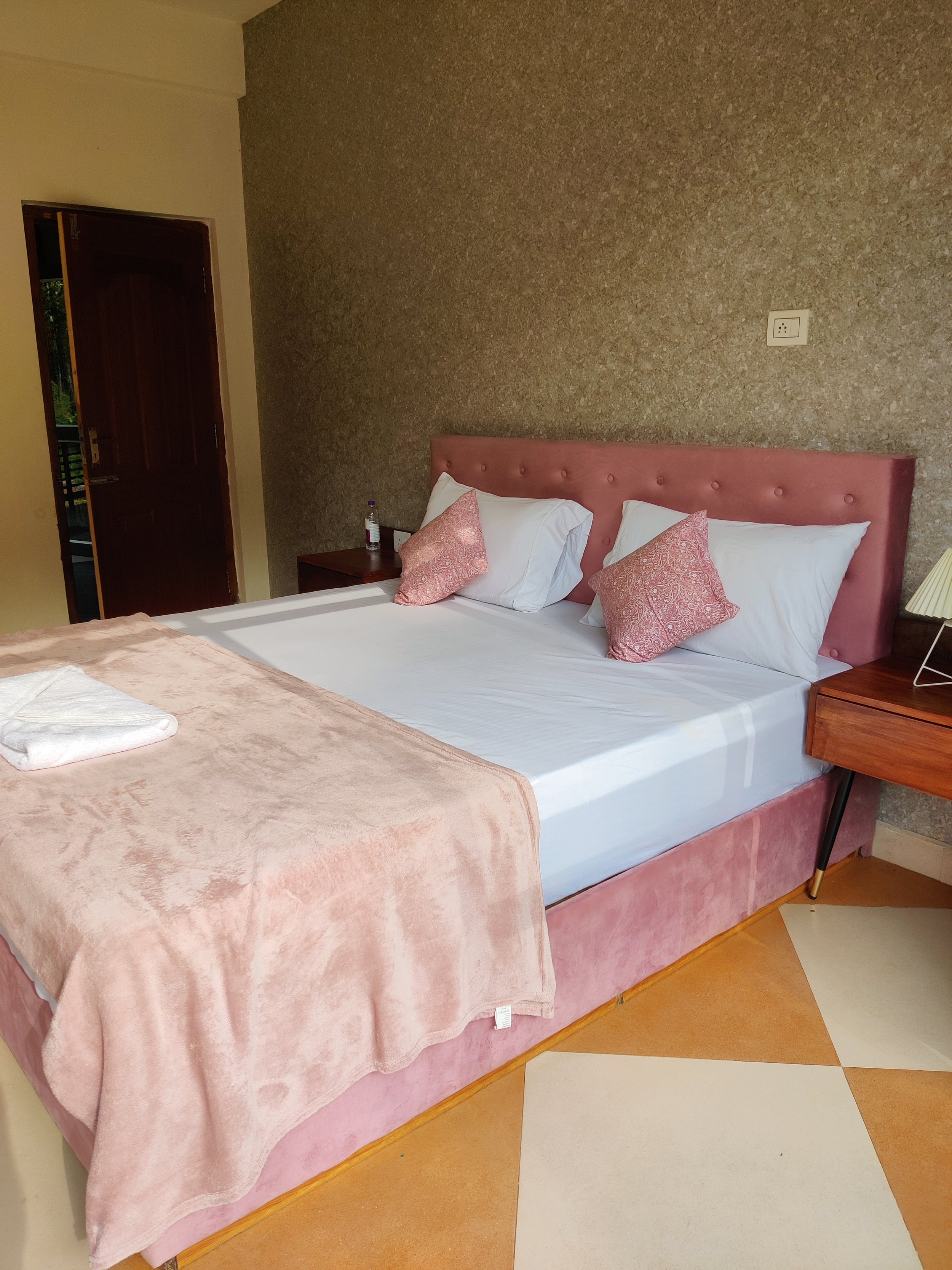 A hotel room with a pink upholstered bed, white sheets, and pink pillows, with a bedside table holding a water bottle, and a wooden nightstand with a lamp. The room has a textured wall behind the bed and a partially open door leading outside.
