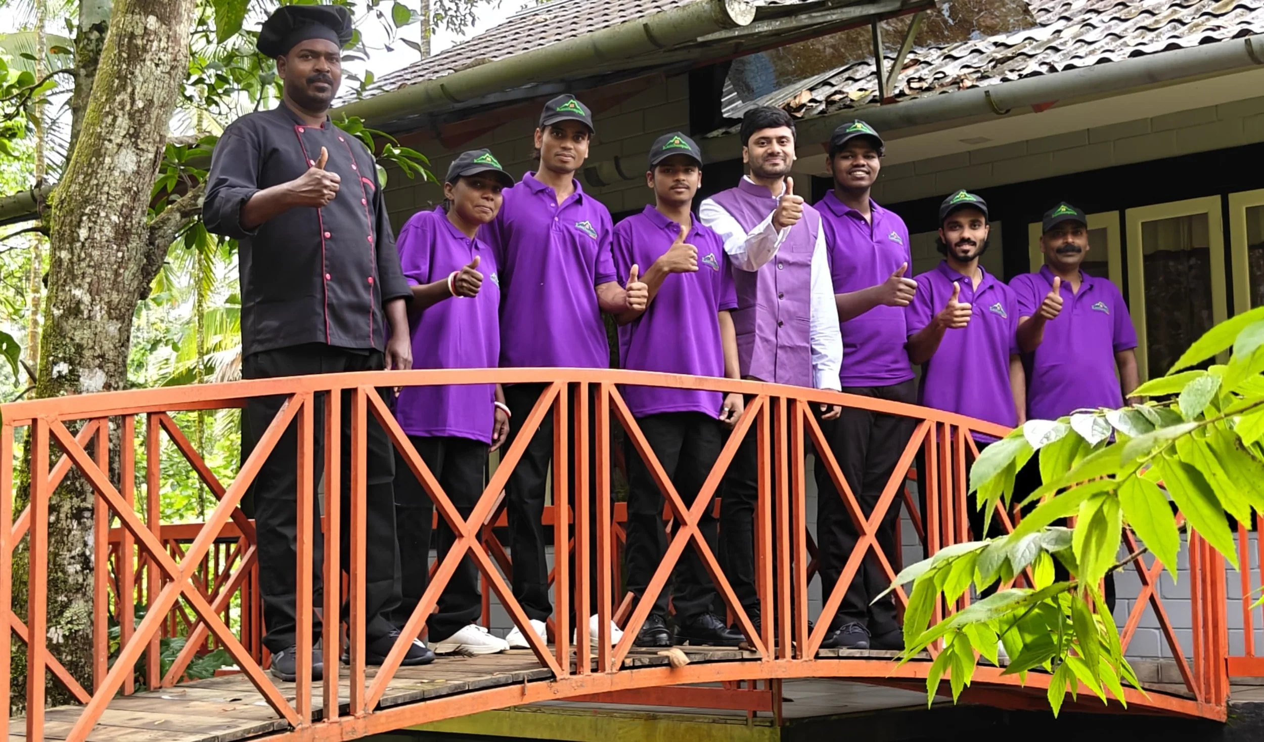 Group of nine people standing on a small orange bridge outside, giving thumbs-up, wearing purple shirts, with a man in a chef's coat on the left, in front of a house and green foliage.