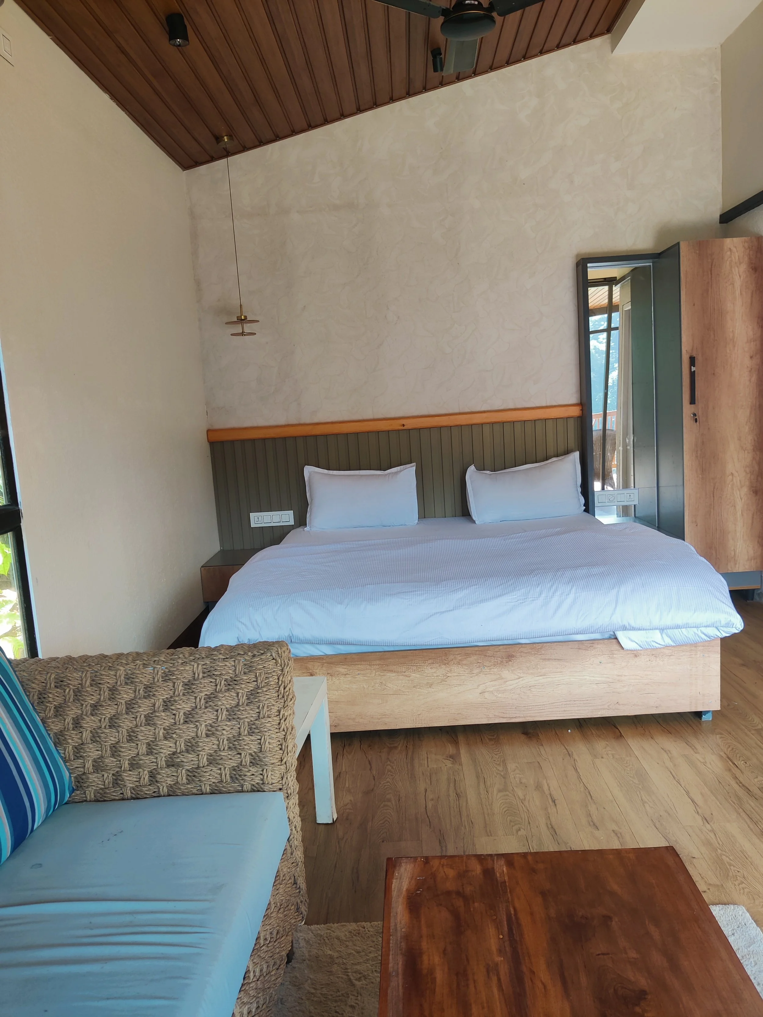 A bedroom with a large bed with white bedding and pillows, a woven armchair with a striped pillow, a wooden coffee table, a tall wardrobe, and a window with visible natural light. The walls are decorated with textured paint and the ceiling is wood-pa