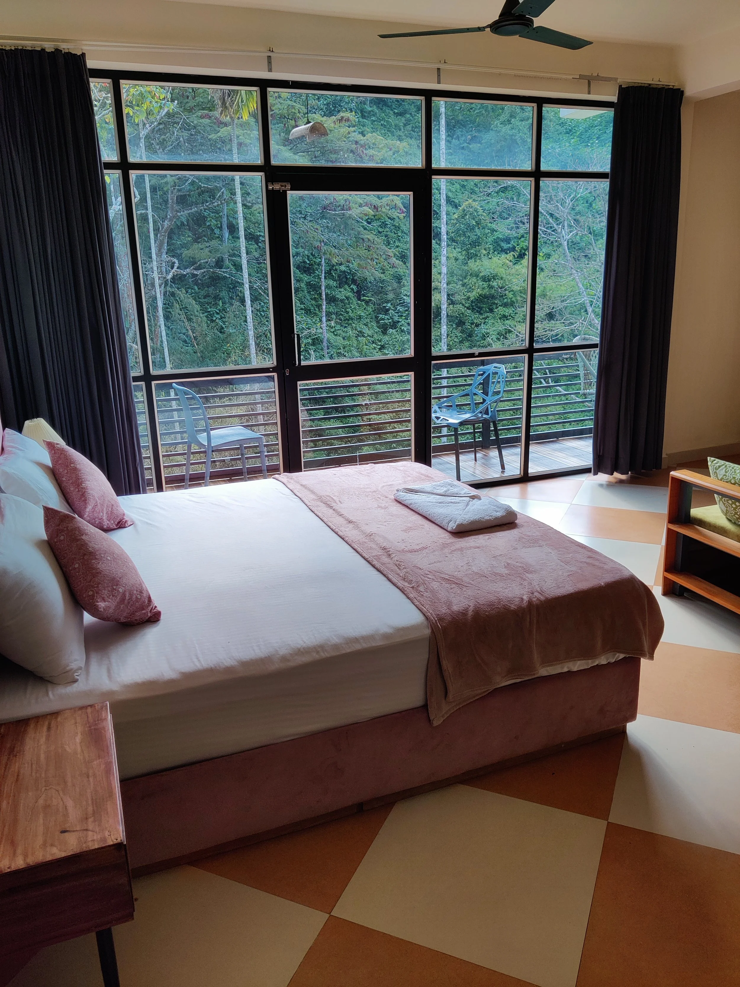 A hotel room with a bed, pink pillows, and a pink blanket, overlooking a balcony with two chairs, surrounded by a dense green forest outside large glass doors and windows.