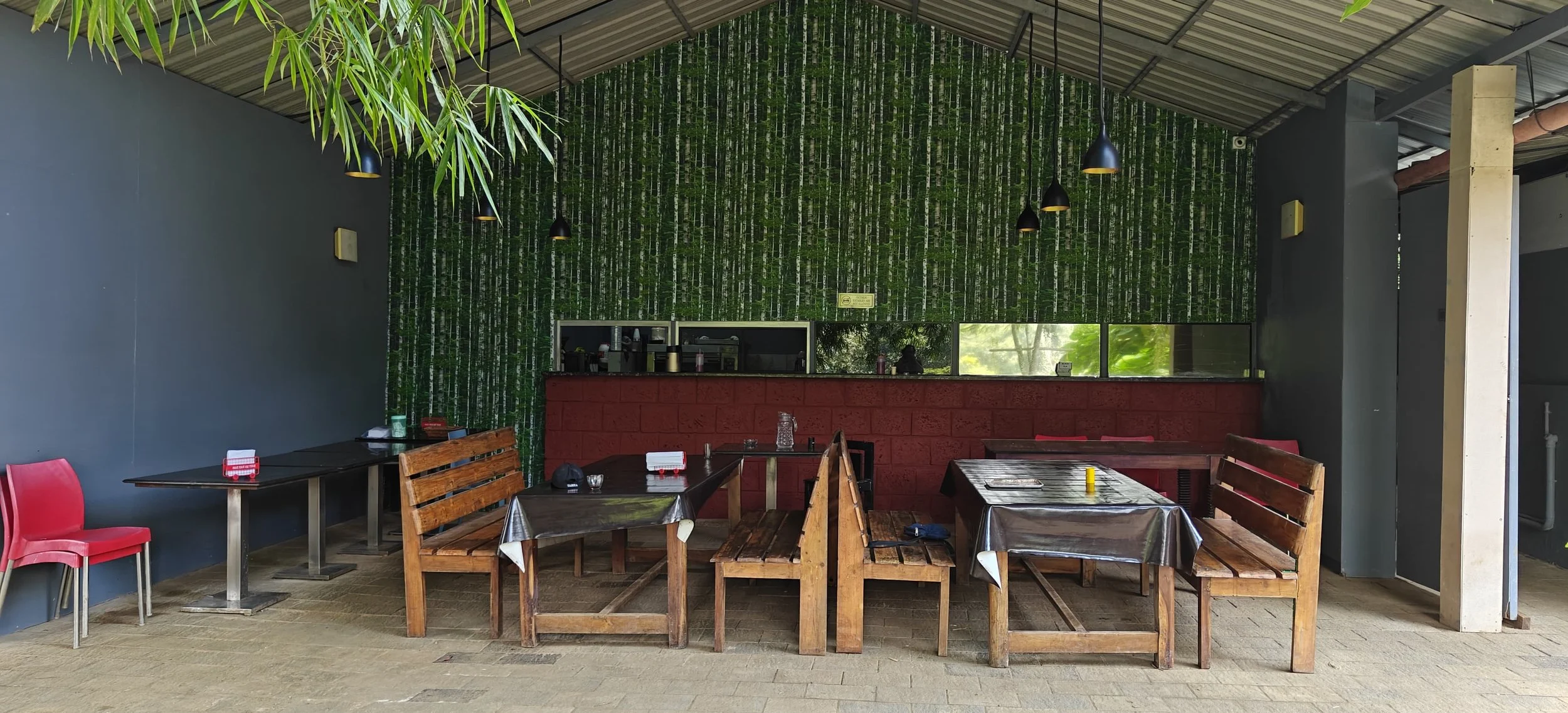 Indoor dining area with wooden benches, tables covered with black and transparent tablecloths, some with condiments and napkins. A red wall is behind the serving window, which is visible in the back. The wall behind the counter features a green bamboo pattern. The ceiling has a metal roof with hanging black pendant lights, and there are some green plants hanging from the ceiling in the foreground.
