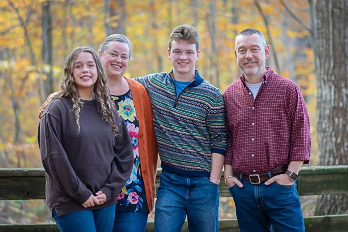 A family of four standing outdoors in front of a wooden fence in a forest with autumn leaves, smiling at the camera.