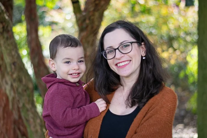 A woman with dark hair, glasses, and a brown sweater holding a young boy wearing a maroon hoodie outside near trees, smiling.