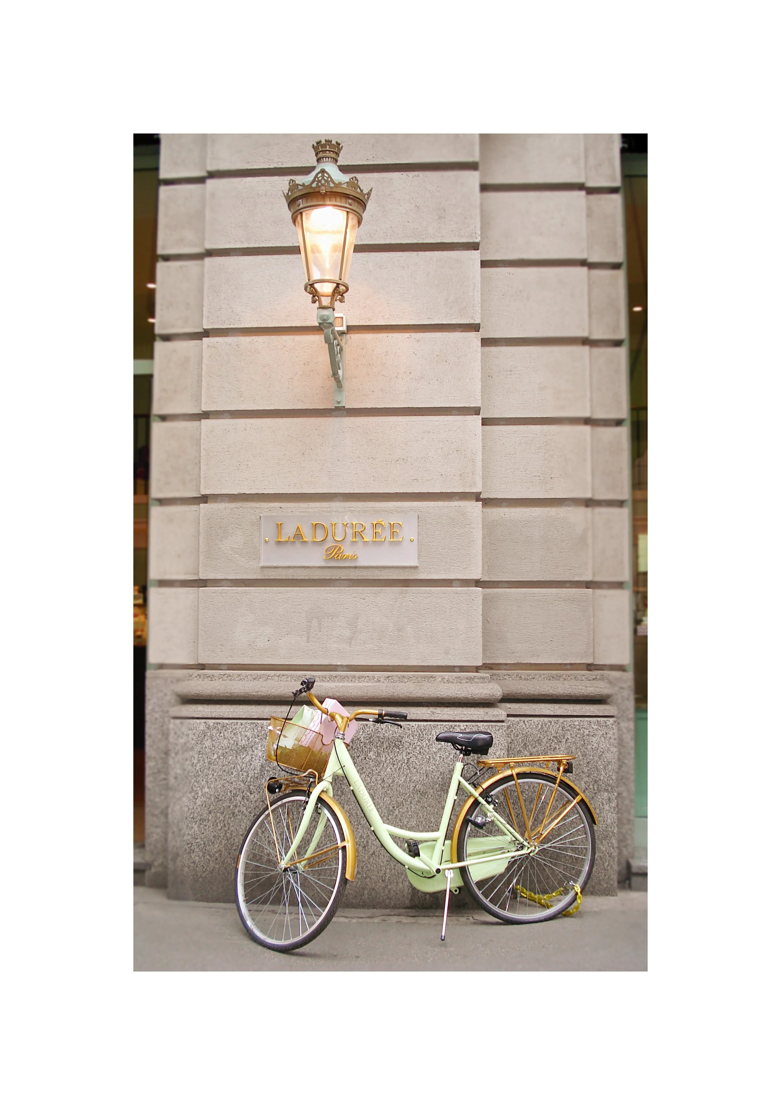 Bicicleta amarela estacionada na calçada com uma parede de prédio ao fundo, uma placa com nome "LADURÉE Paris" em uma parede e uma lanterna de rua na parede ao lado.