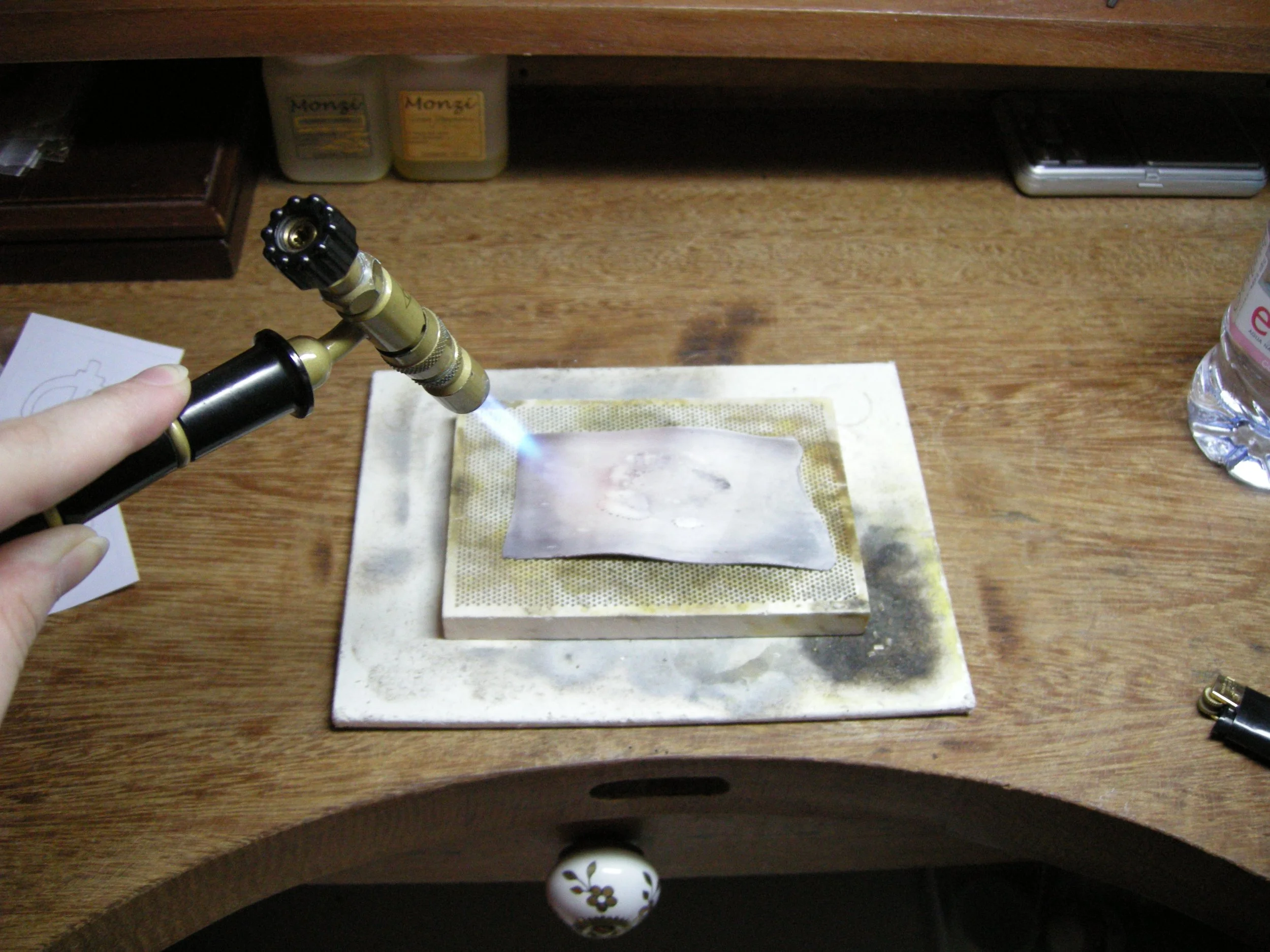 Imagem de uma mesa de madeira com uma pessoa segurando um maçarico de ponta preta, segurando uma bandeja de metal, com álcool gel, um celular do lado direito e duas garrafas de pasta deSetting Monzi no fundo.