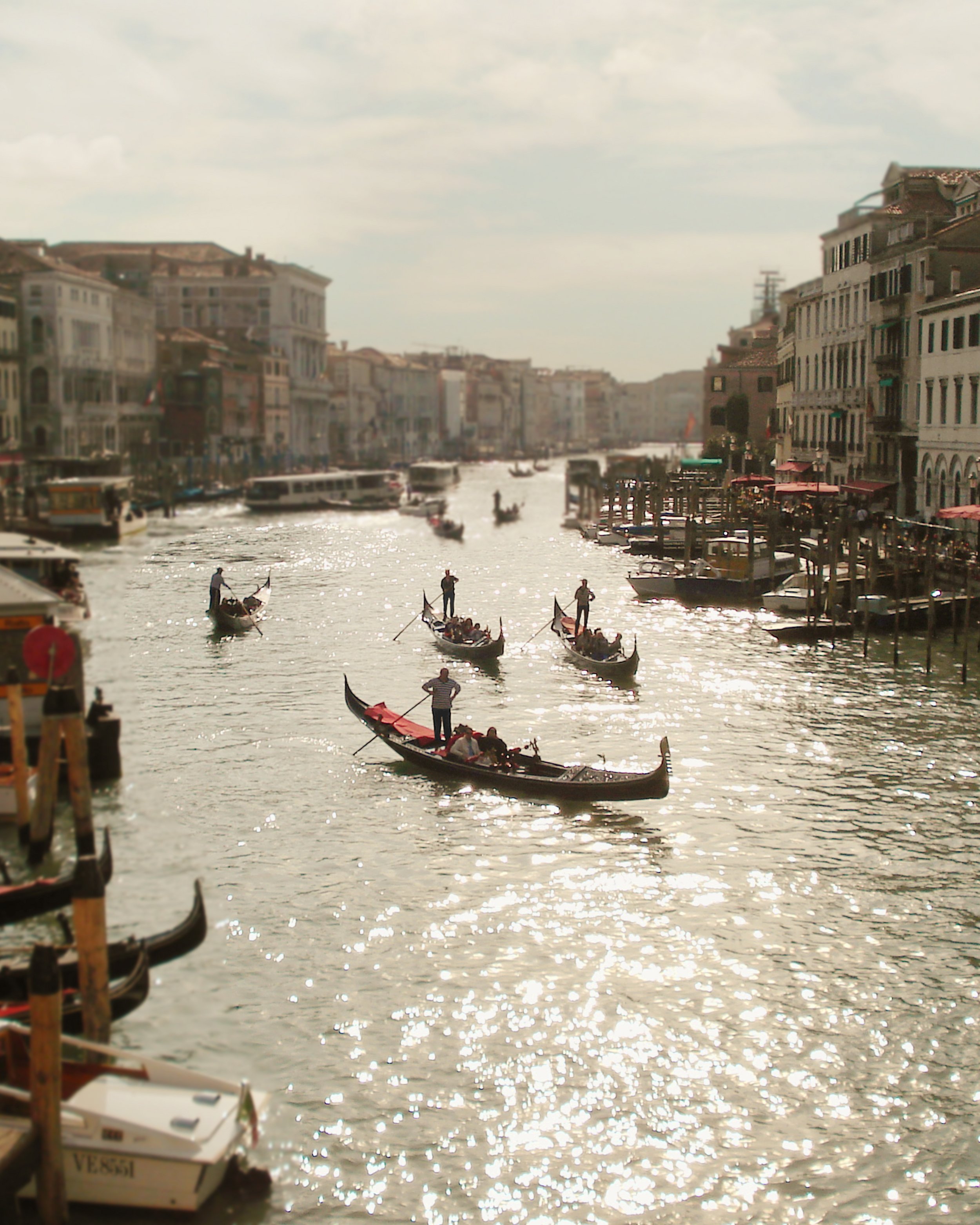 Imagem de um canal de Veneza com diversas gôndolas e barcos navegando, ao lado de prédios históricos e cafés ao ar livre ao longo do rio.