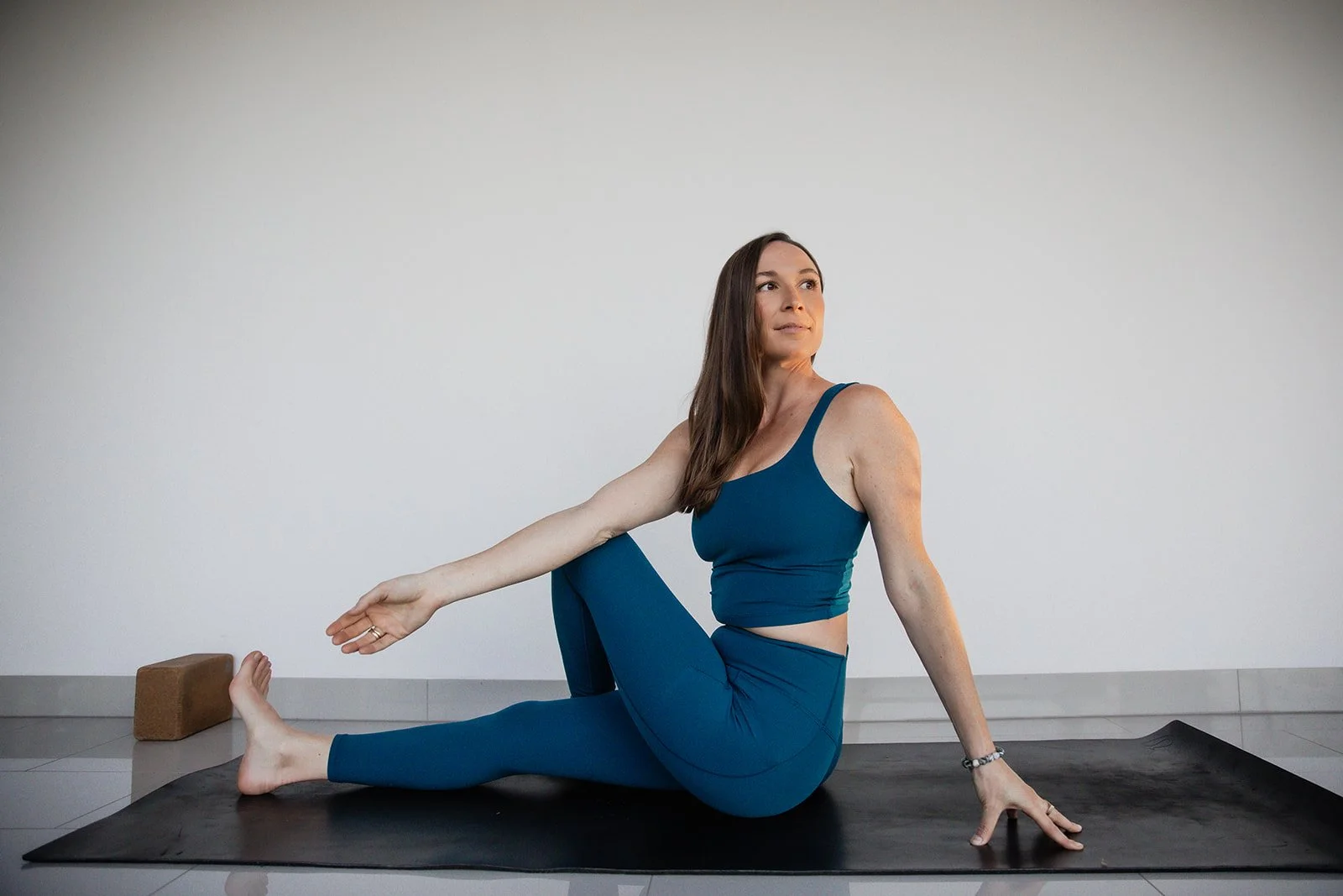 Woman practicing yoga seated on black mat, with yoga block nearby, against plain white wall.