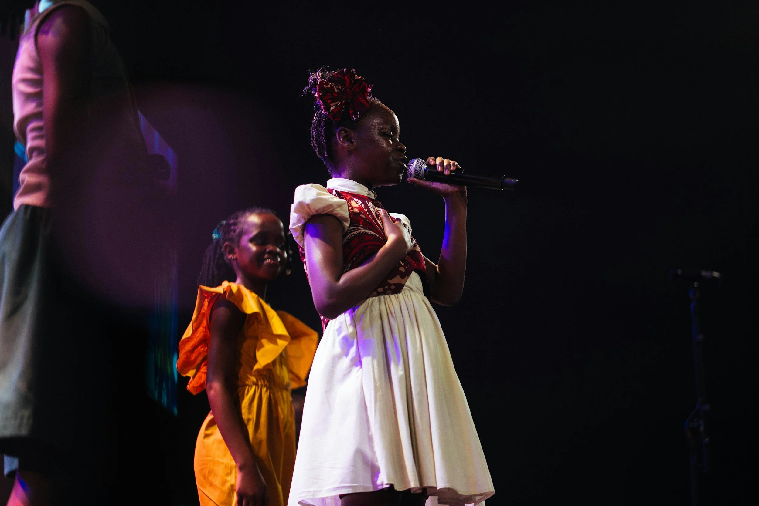 A woman in a dress holding a microphone on stage, with a young girl in a yellow dress smiling beside her, against a dark background.