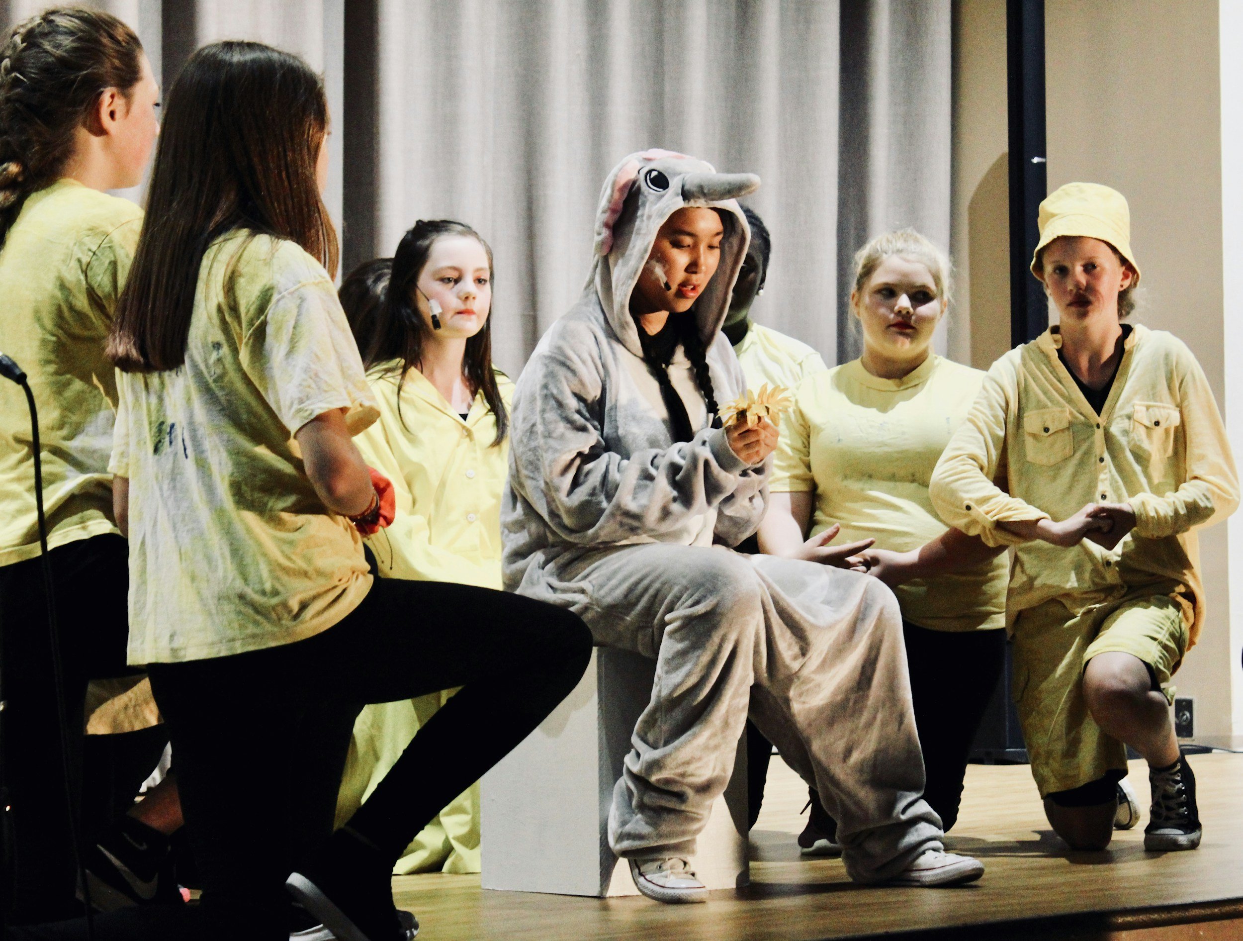 Group of children on stage during a school play, some dressed in beige costumes and one in a gray animal onesie with an elephant or elephant-like head, others in yellow clothing. One child in pajamas is seated and holding a leaf.
