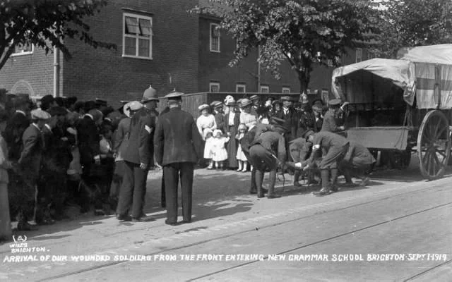 A line of people, including children and adults, waiting to enter New Grammar School in Brighton, September 1919, as wounded soldiers arrive in a horse-drawn wagon.