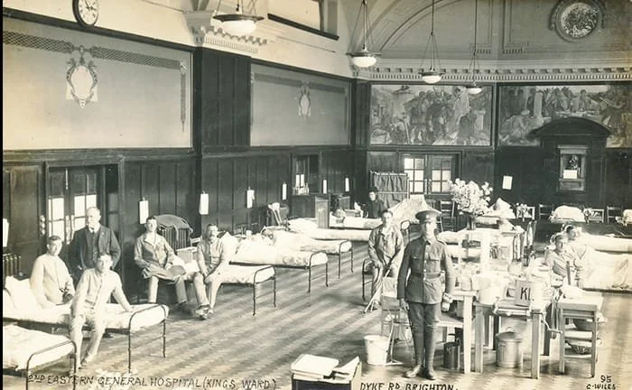 Black and white photograph of a hospital ward with beds and several patients. A nurse or orderly is standing near a table with medical supplies. The room has wooden paneling and large murals on the wall.