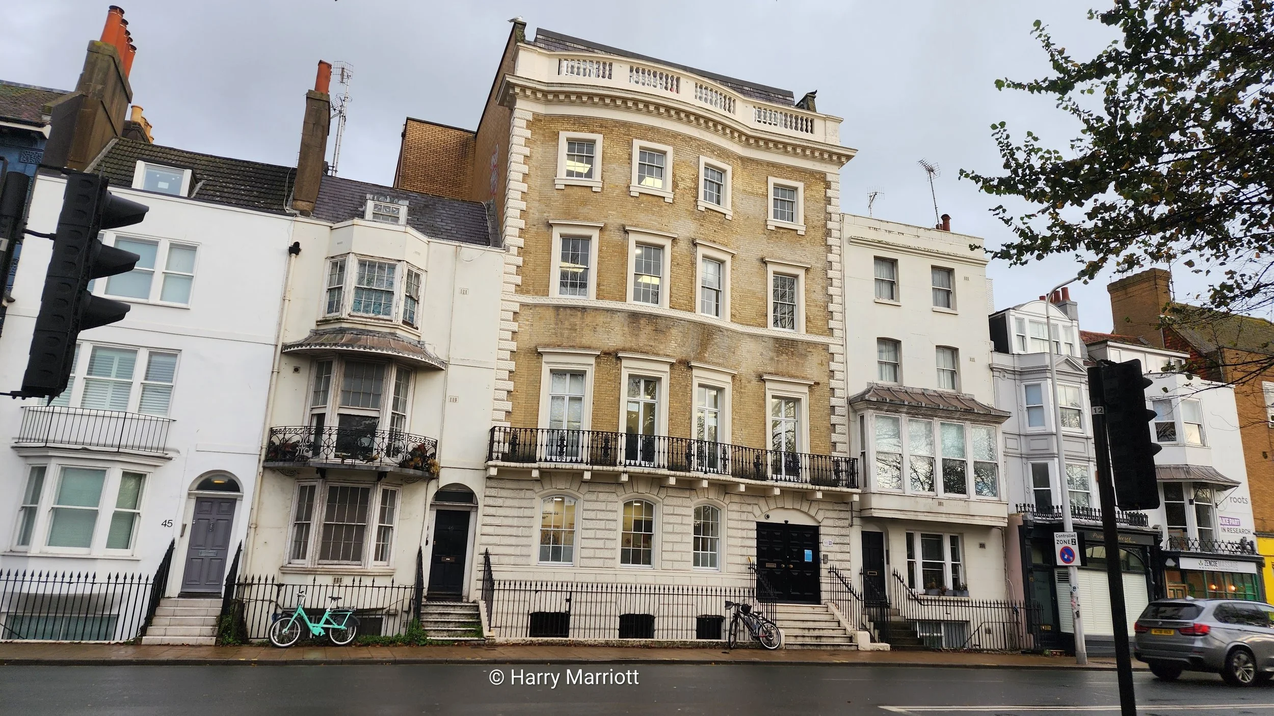 Row of Victorian-style multi-story buildings on a city street, with a bicycle and a car parked outside, and traffic lights on either side.