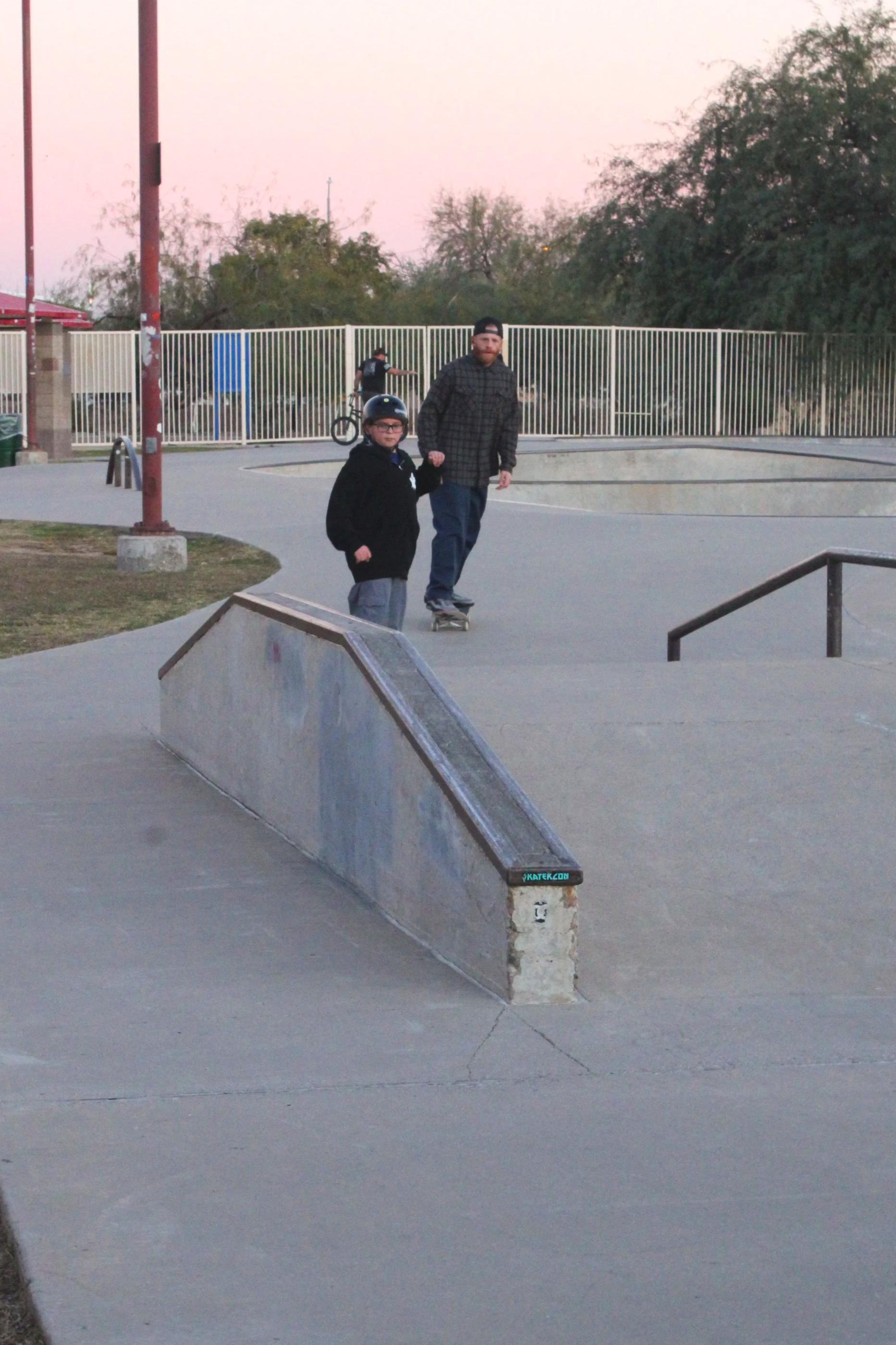 A skate park with two skateboarders, one an adult and the other a child, riding near a small ramp during sunset with trees and a fence in the background.