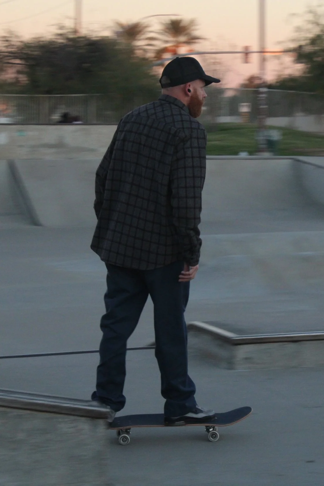 A man with a beard and a black cap skateboarding at a skate park during sunset.