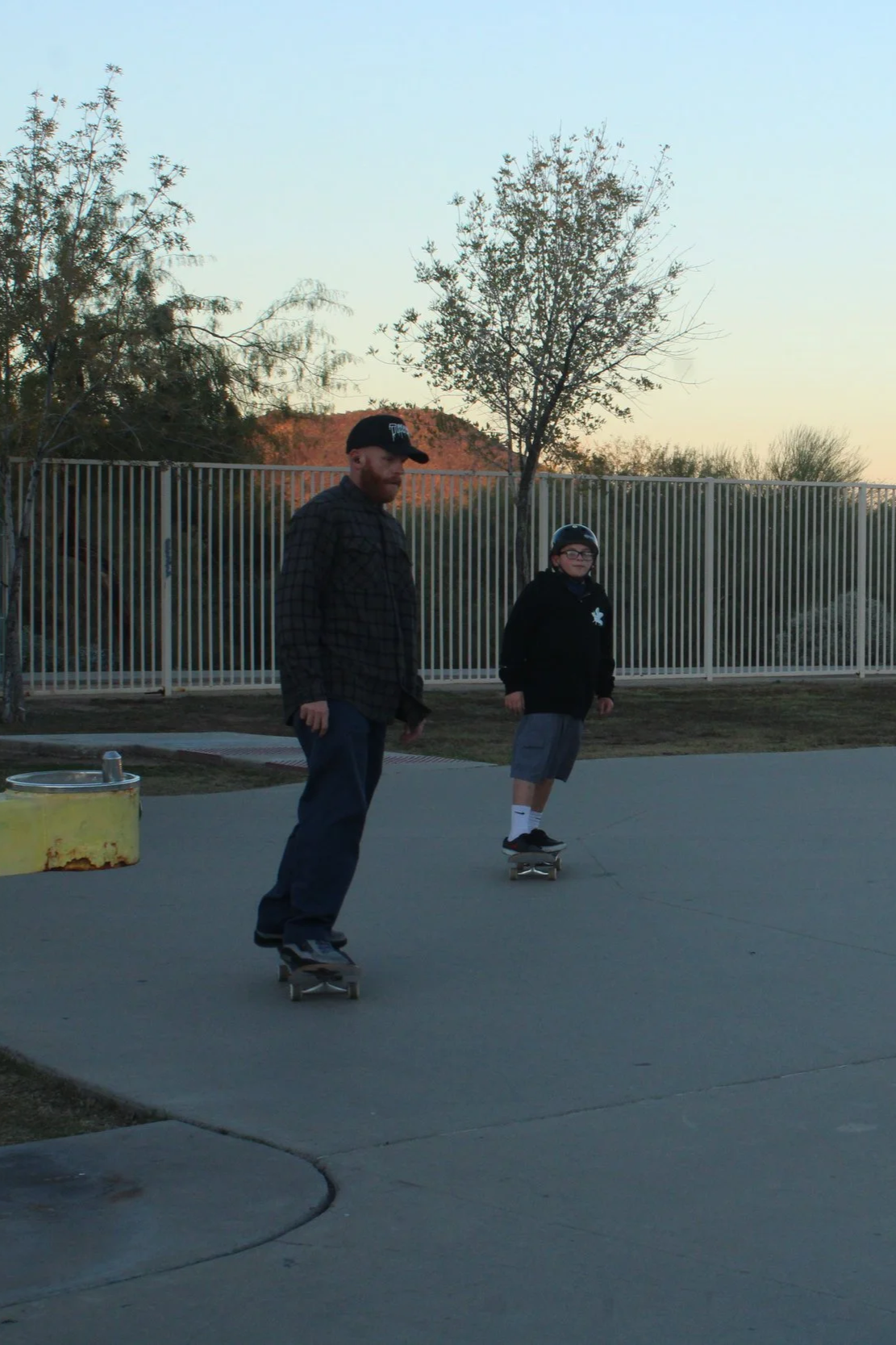 A man and a boy skateboarding outdoors during sunset with a fence and trees in the background.
