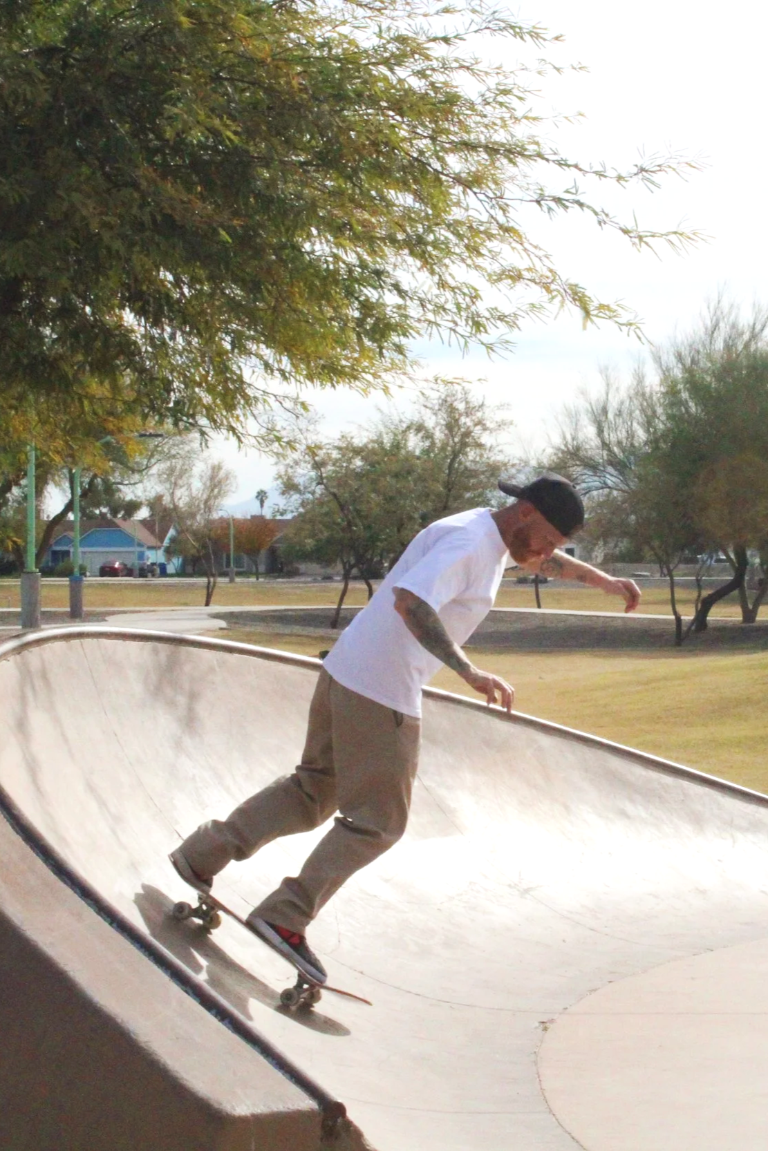 Man skateboarding in a skatepark bowl during daytime with trees and a park in the background.