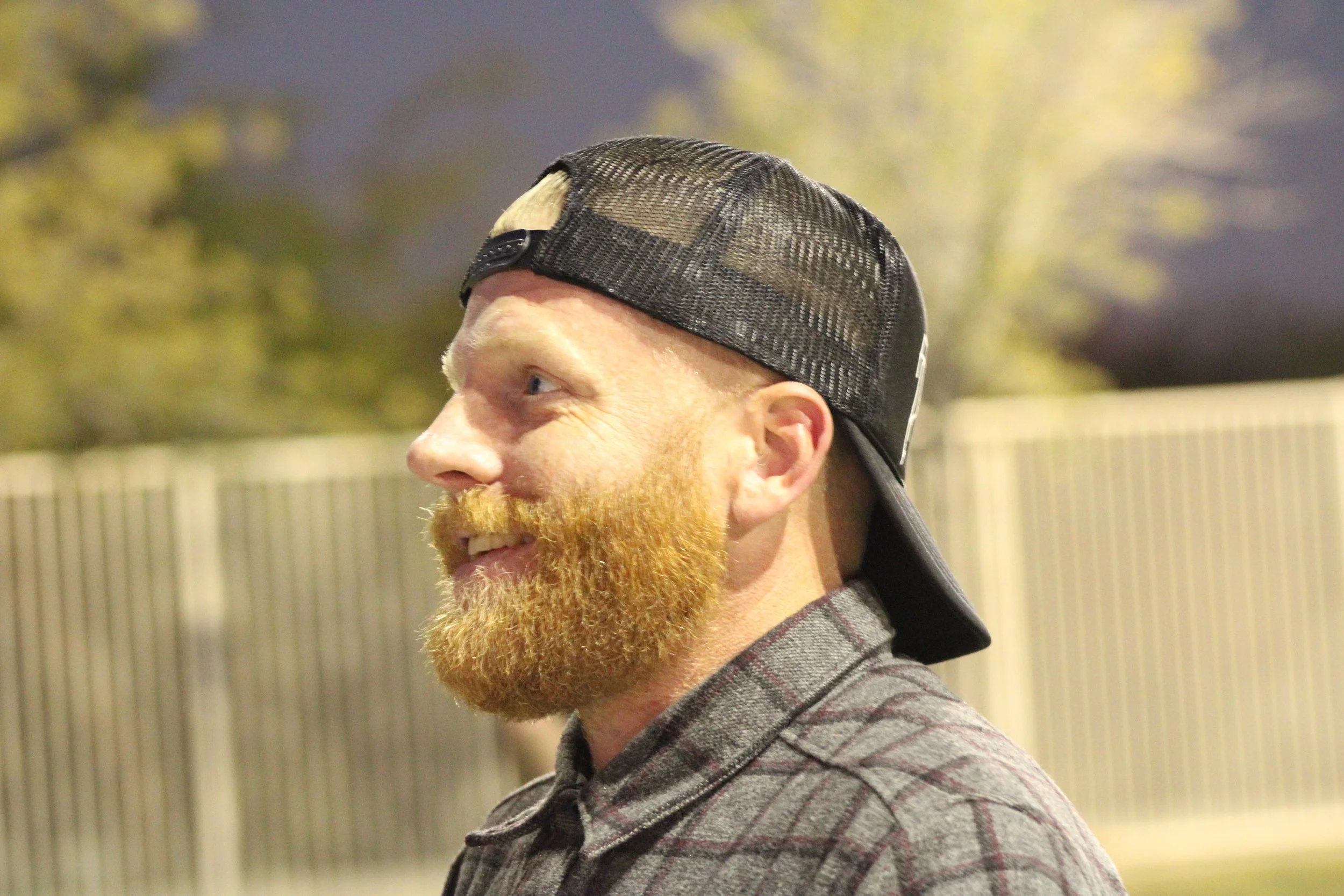 A man with a red beard wearing a black baseball cap backwards and a plaid shirt, smiling outdoors during the evening or night.