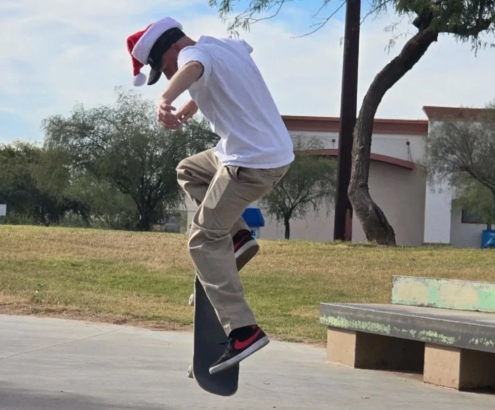 A person wearing a Santa hat, white shirt, and khaki pants performs a skateboard trick at a park.