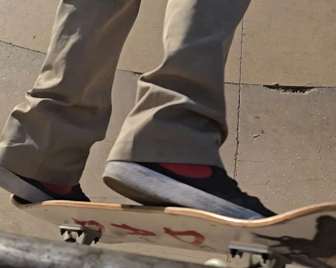 A person wearing beige pants and black sneakers riding a skateboard on a concrete sidewalk.