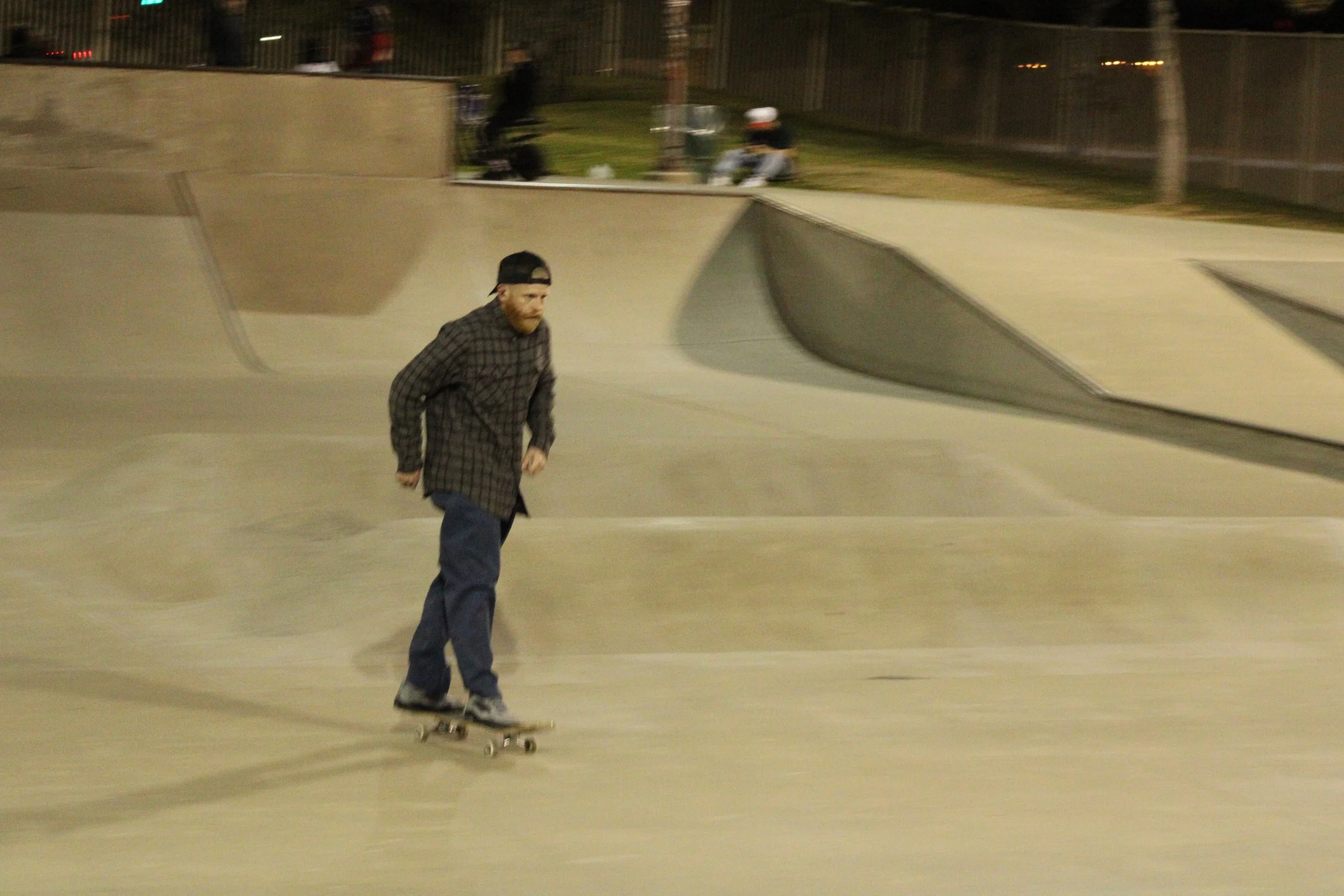 A man with a beard wearing a checkered shirt, dark pants, and a backward baseball cap skateboarding at a concrete skate park at night.