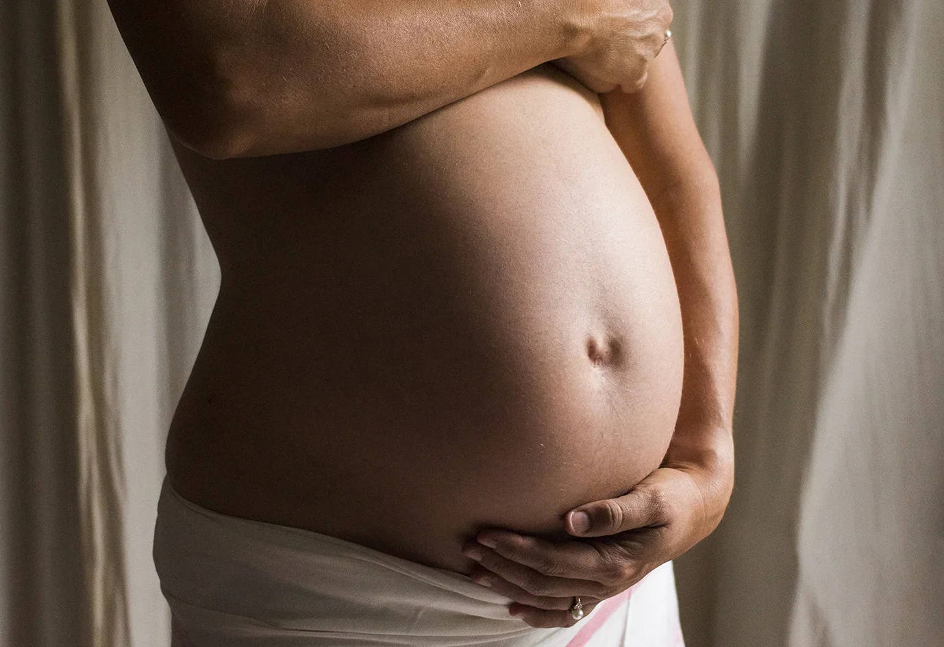 A pregnant woman gently cradling her belly with one hand, standing in front of a neutral background.