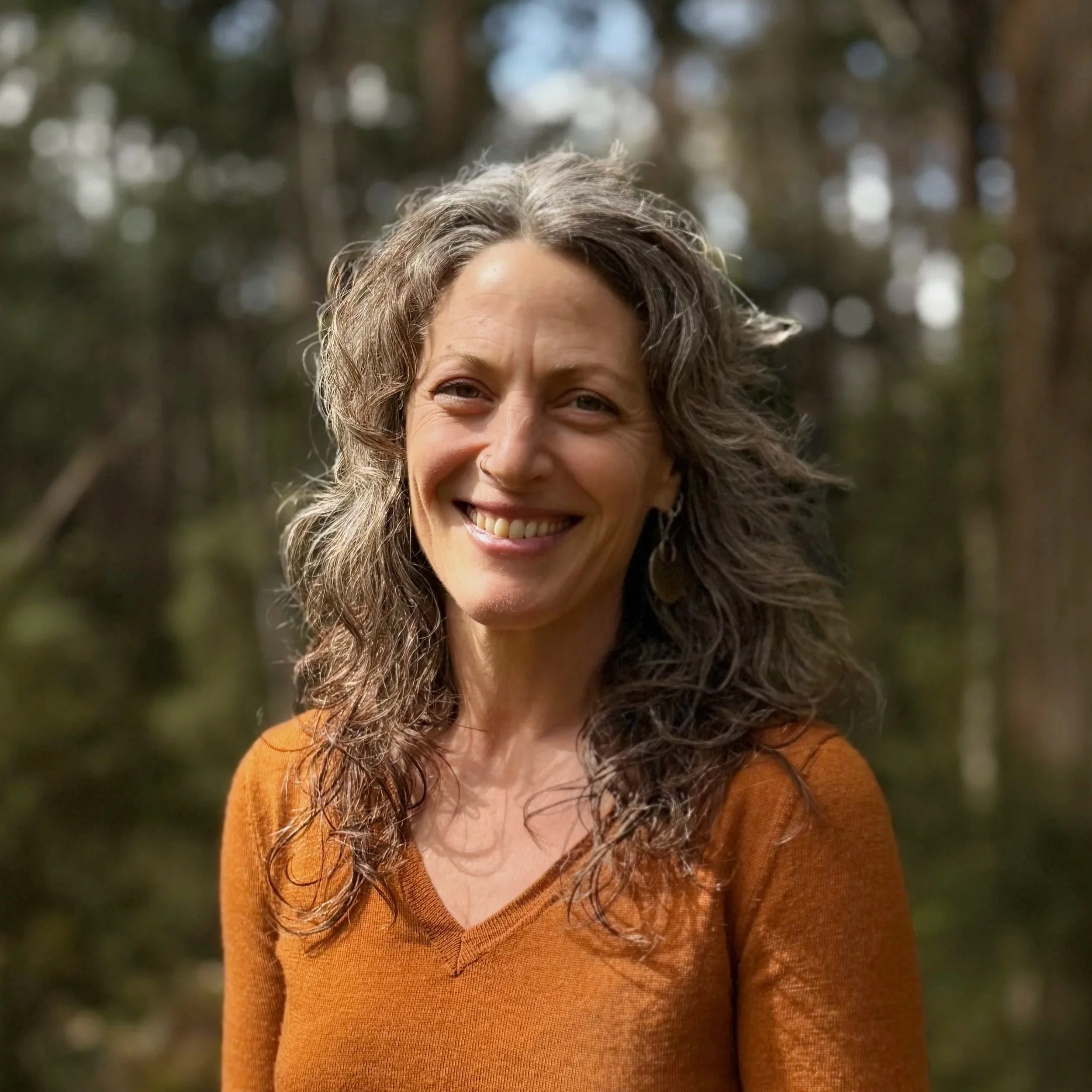 A smiling woman with wavy, gray hair wearing a rust-colored sweater outdoors in a forested area.