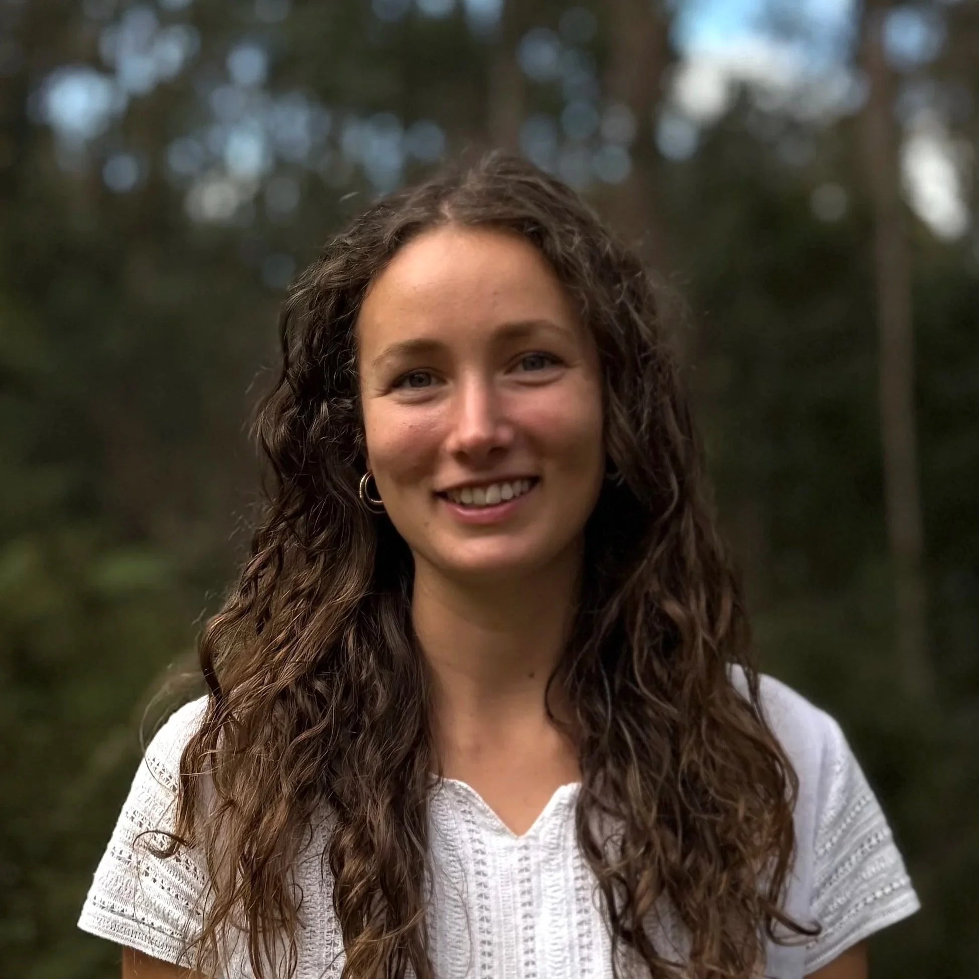 Young woman with long, curly brown hair smiling outdoors with a blurred forest background.