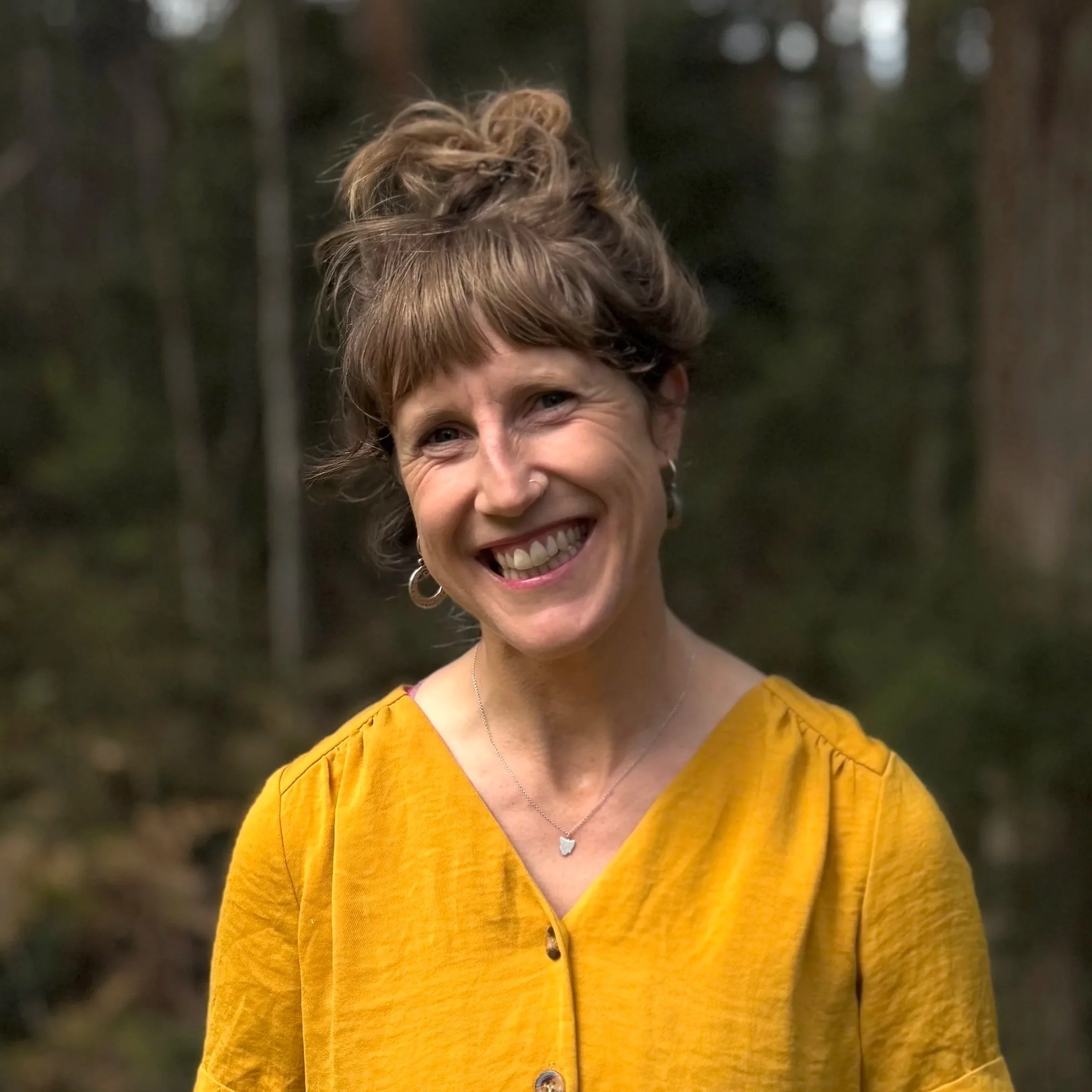 A woman with short, curly brown hair smiling outdoors in a forest, wearing a yellow button-up top, earrings, and a necklace.