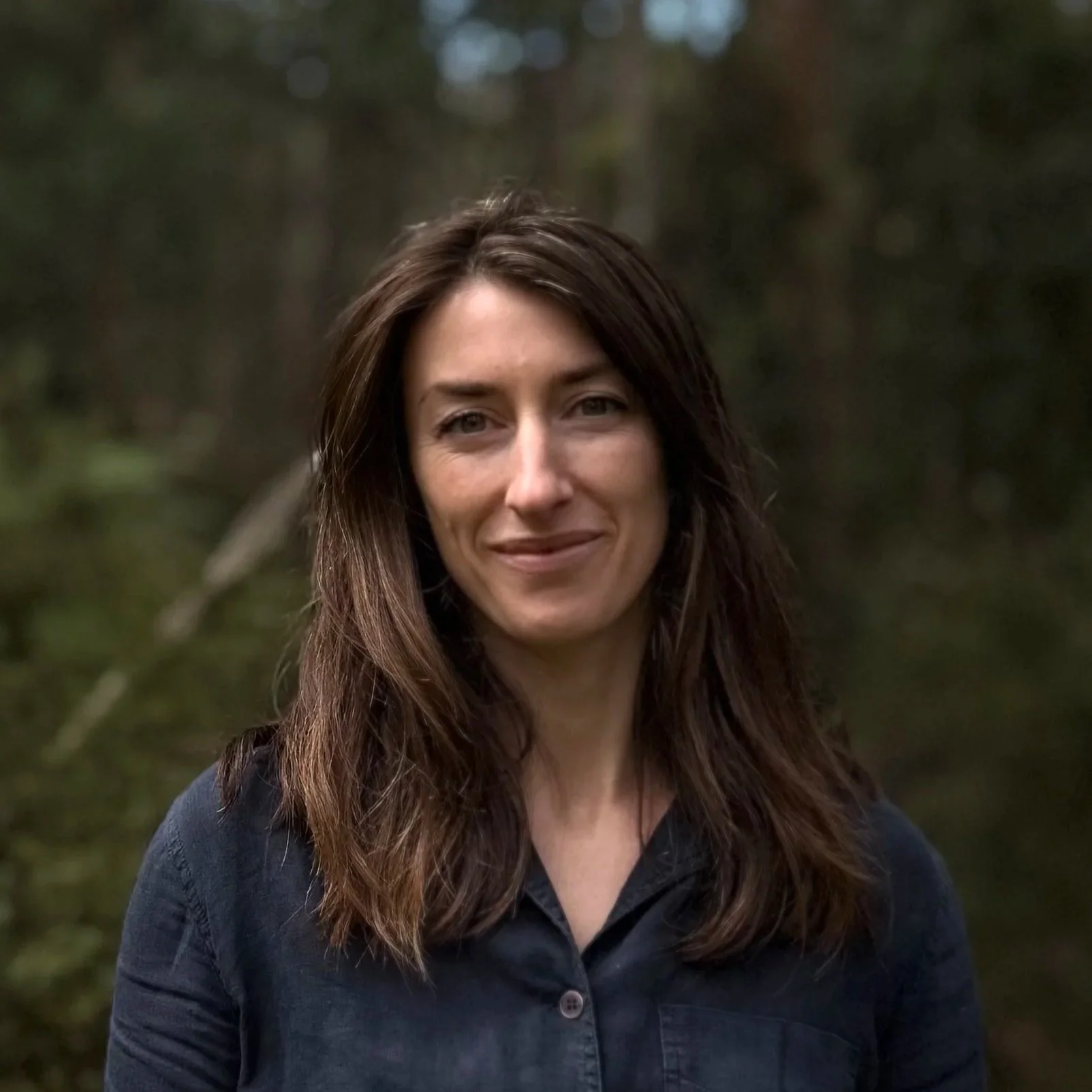A woman with long brown hair and a slight smile standing outdoors in a forested area.