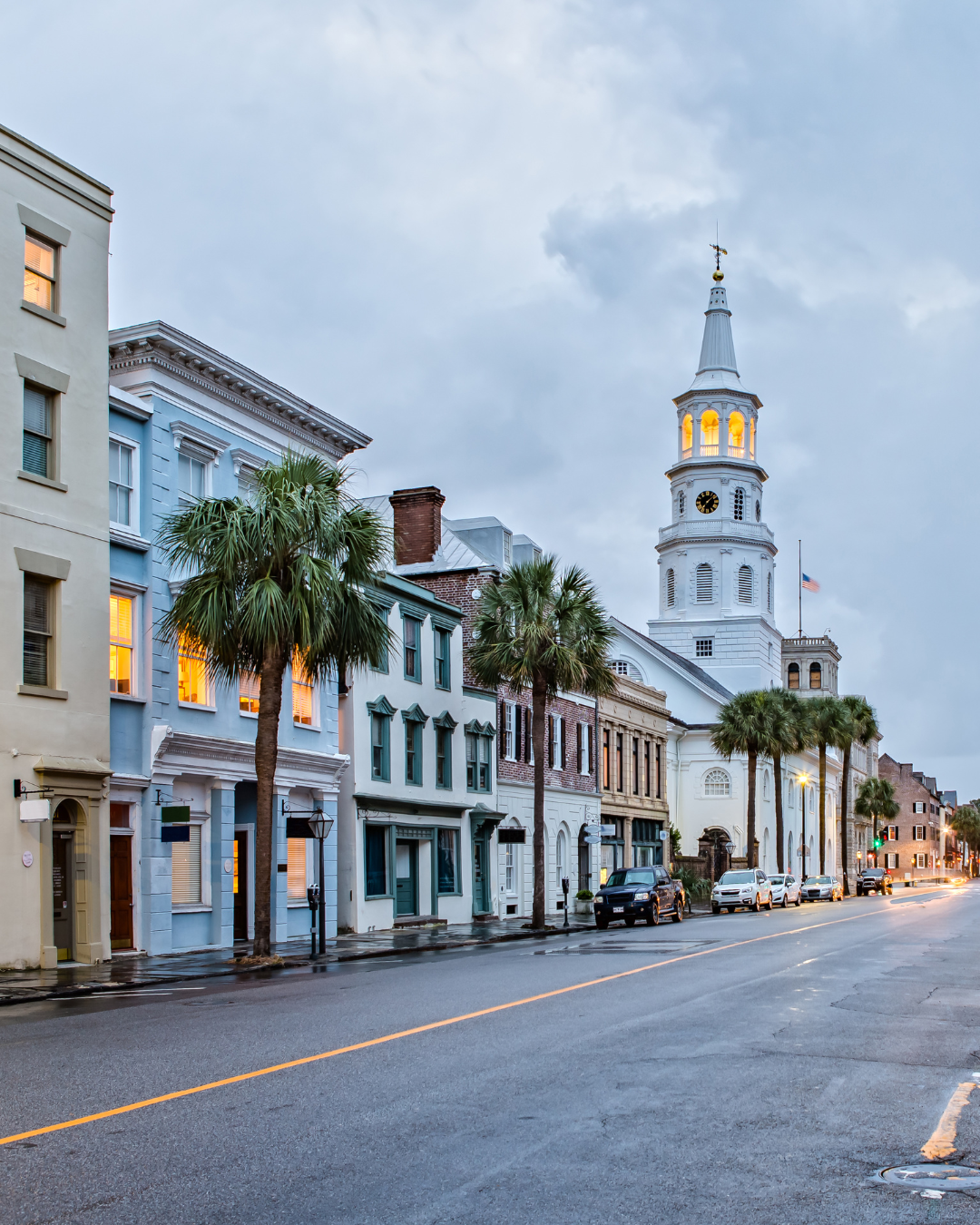 Street view of historic buildings with palm trees, cloudy sky, and a prominent white church with a steeple and lit windows in Charleston, South Carolina.