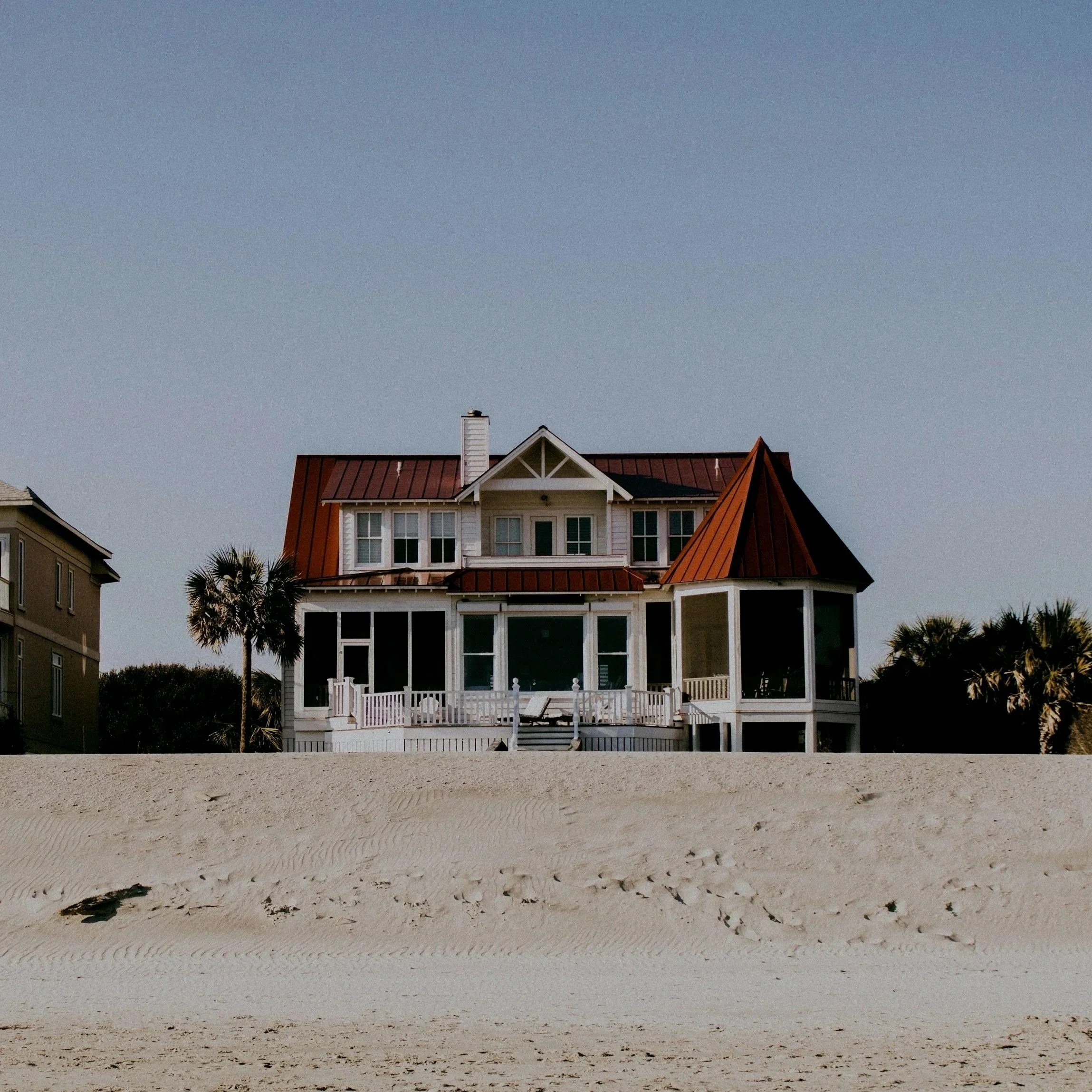 A large beach house with a red metal roof, white walls, and large windows, situated on a sandy beach with palm trees and a clear sky in the background.