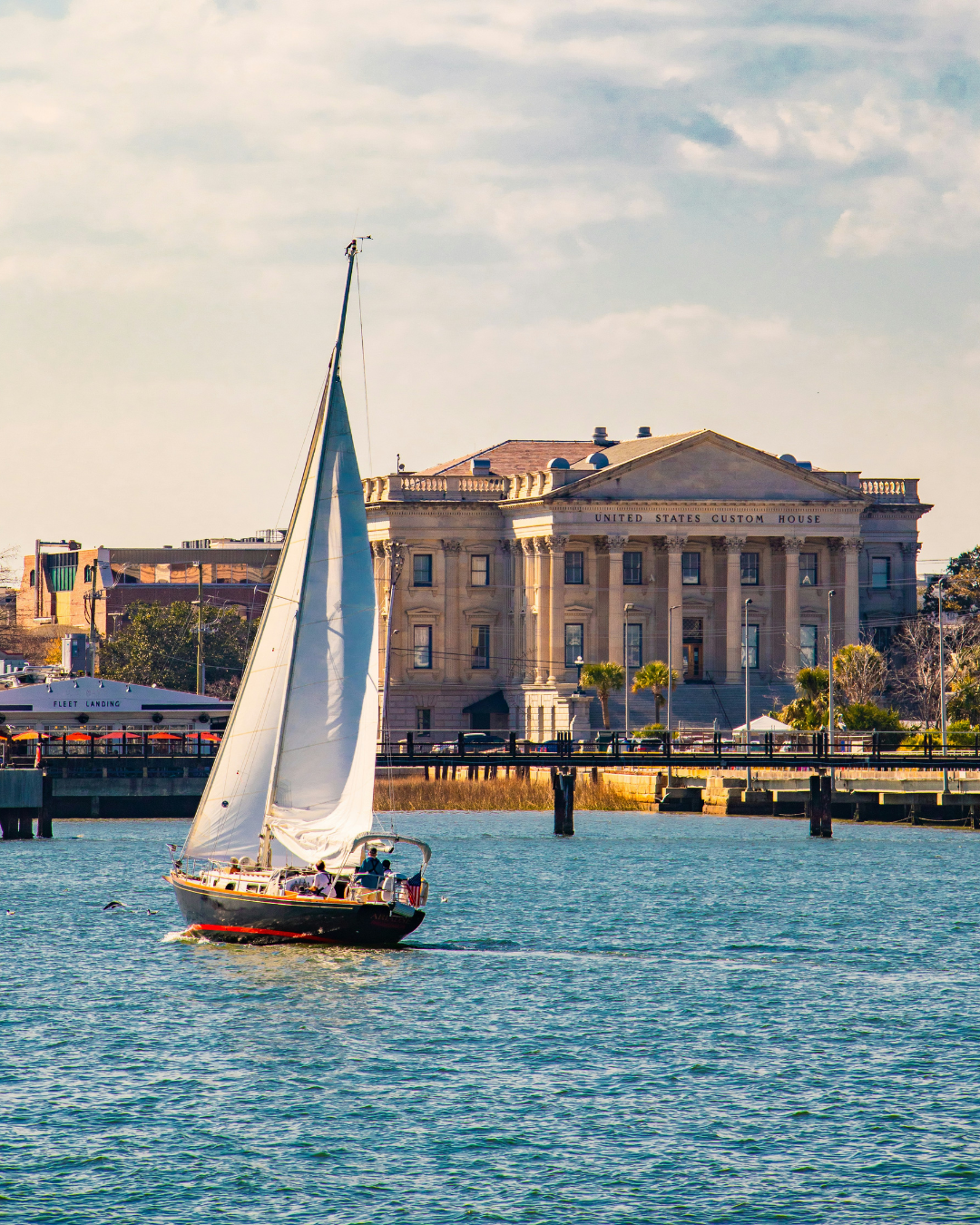 Sailboat sailing on water in front of an historic government building with columns, labeled 'United States Custom House', and trees along the shoreline.