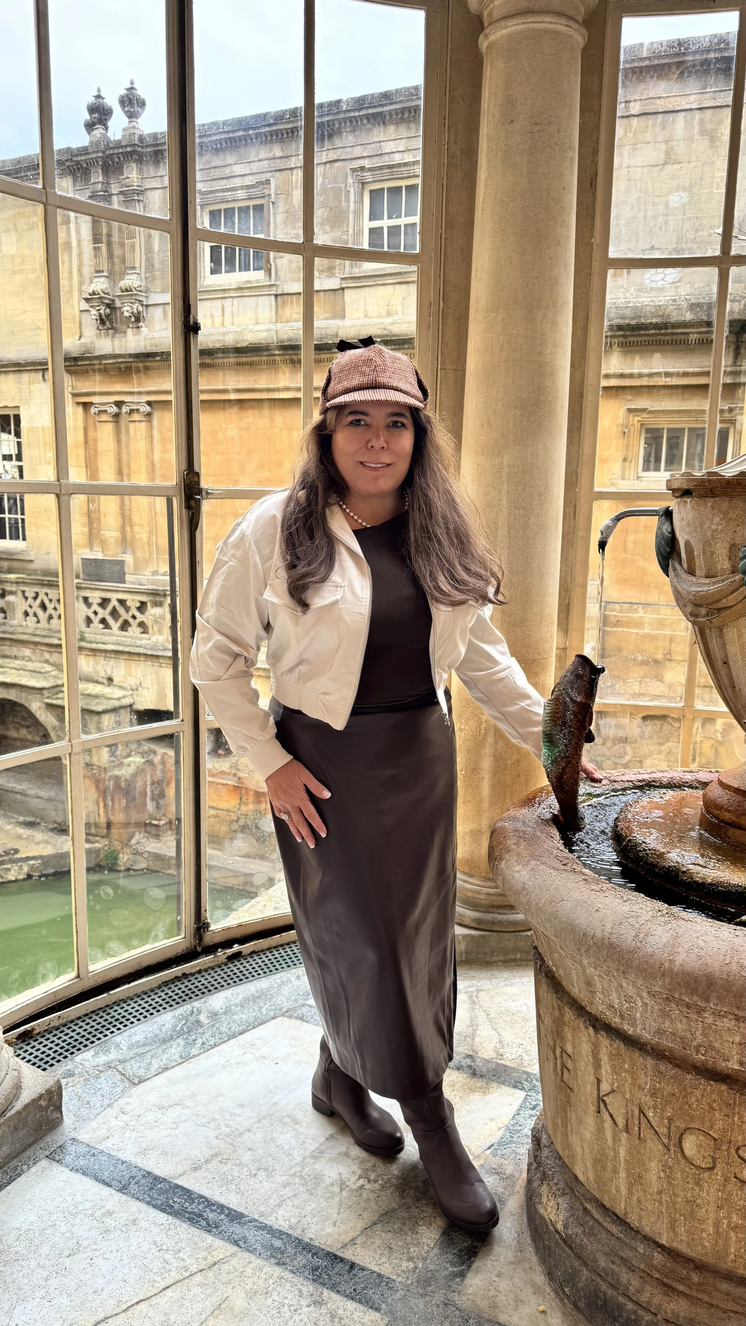 Woman standing indoors near a fountain with a fish sculpture, wearing a pink cap, a white jacket, a black top, a long skirt, and boots, with historic building windows in the background.