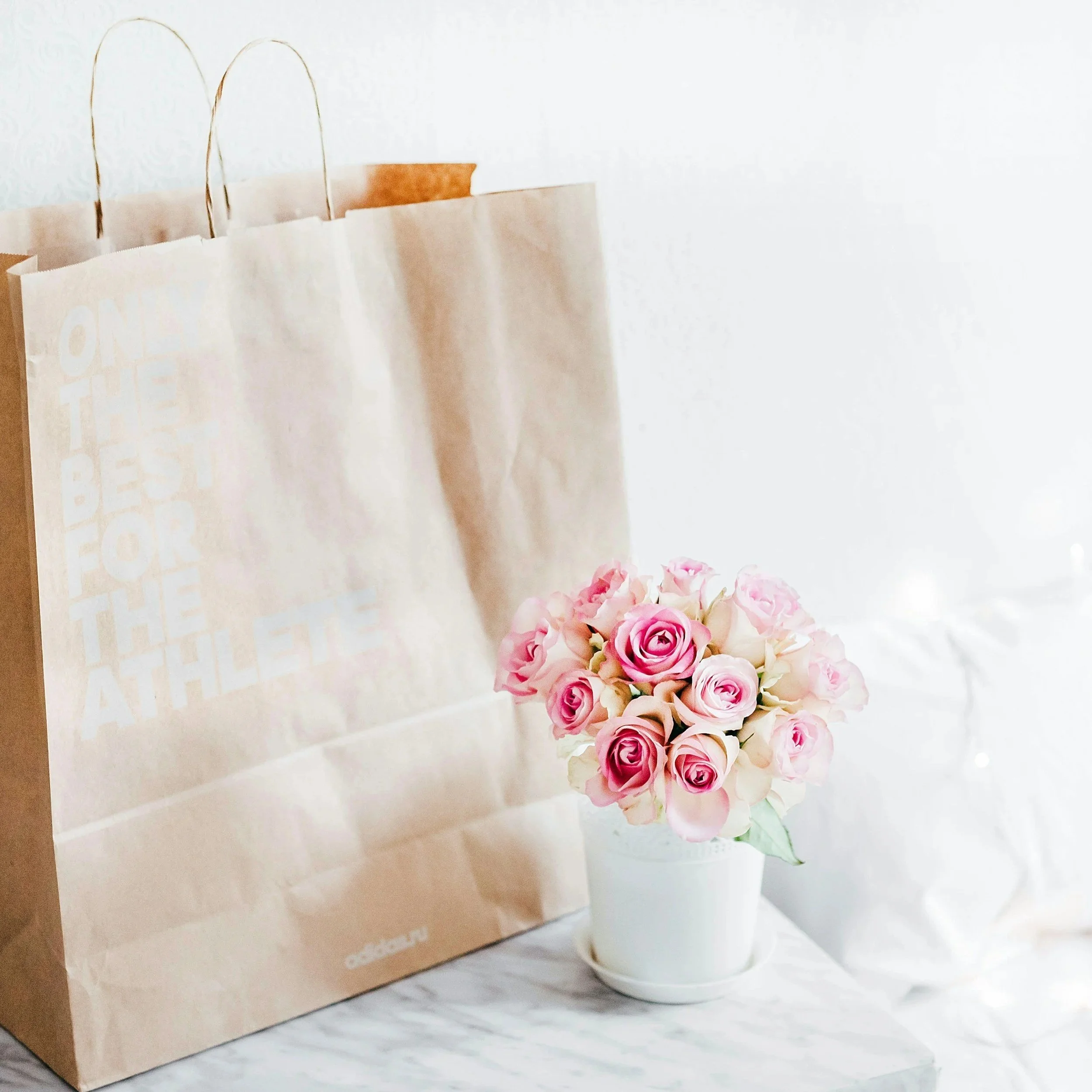 A bouquet of pink and white roses in a white vase on a marble surface, with a beige shopping bag in the background.