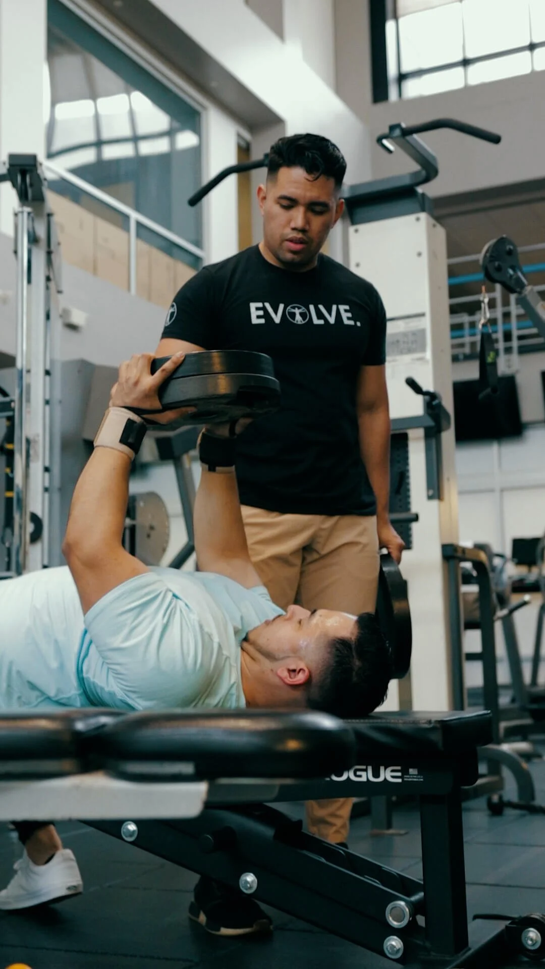 A man is lying on a workout bench doing a dumbbell chest press with a dumbbell in each hand, while a personal trainer stands beside him in a gym, spotting him.