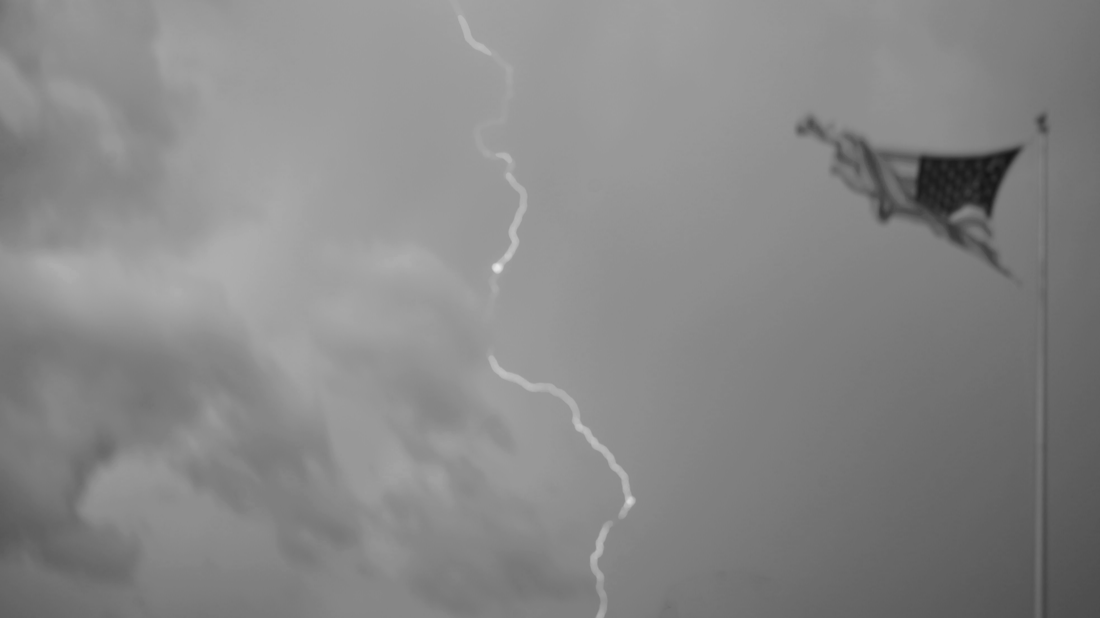 Lightning bolt in a cloudy sky with an American flag in the background.