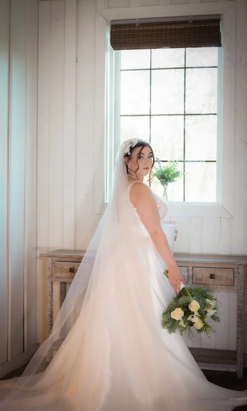 Bride in a white wedding gown with veil holding a bouquet of white roses and greenery, standing indoors in front of a large window with natural light
