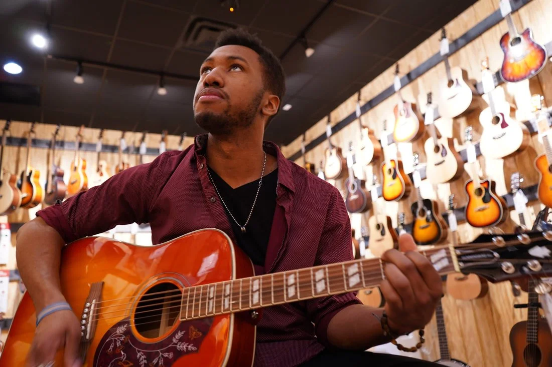 A young man playing an acoustic guitar inside a music store with guitars hanging on the wooden wall behind him.