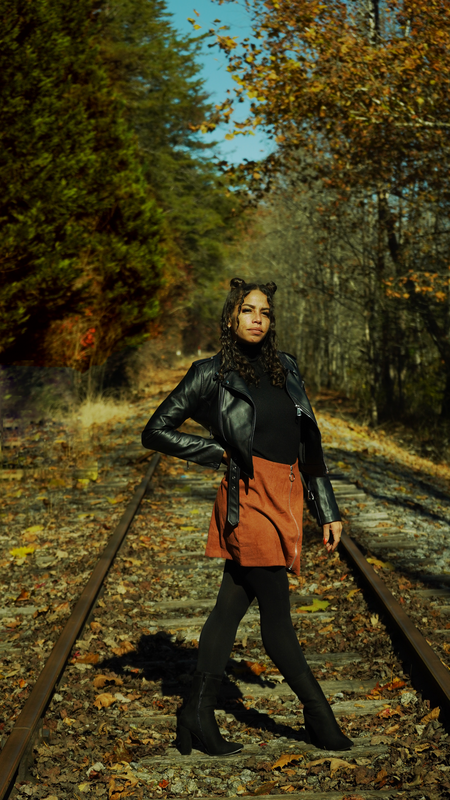 A young woman standing on abandoned train tracks in a forest during fall, wearing a black leather jacket, black turtleneck, brown skirt, black leggings, and black boots.