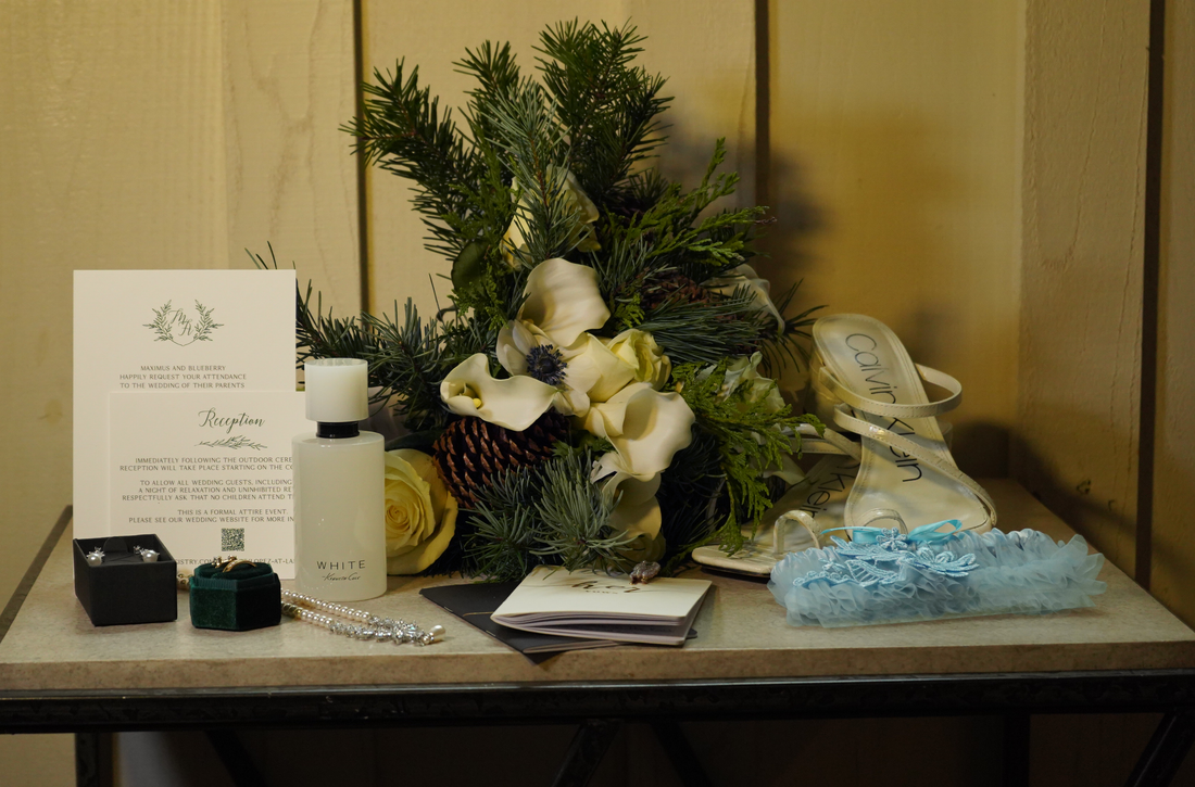 Wedding guest table with a floral arrangement, wedding shoes, a bottle of perfume, jewelry, an invitation card, and a small blue garter.