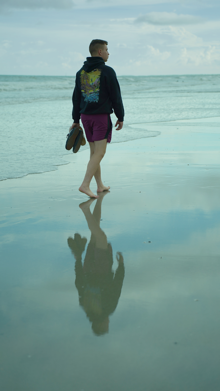Person walking barefoot on a wet beach shoreline holding shoes, with ocean and cloudy sky in the background, and reflection on the wet sand.