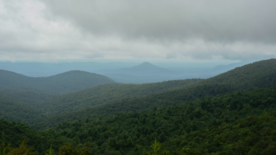Landscape view of green, forested mountains under a cloudy sky, with distant peaks on the horizon.