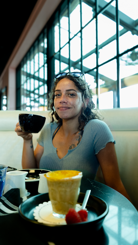 A young woman with curly hair and a gray shirt sitting in a restaurant, holding a coffee cup, and a latte on the table, and large windows in the background.