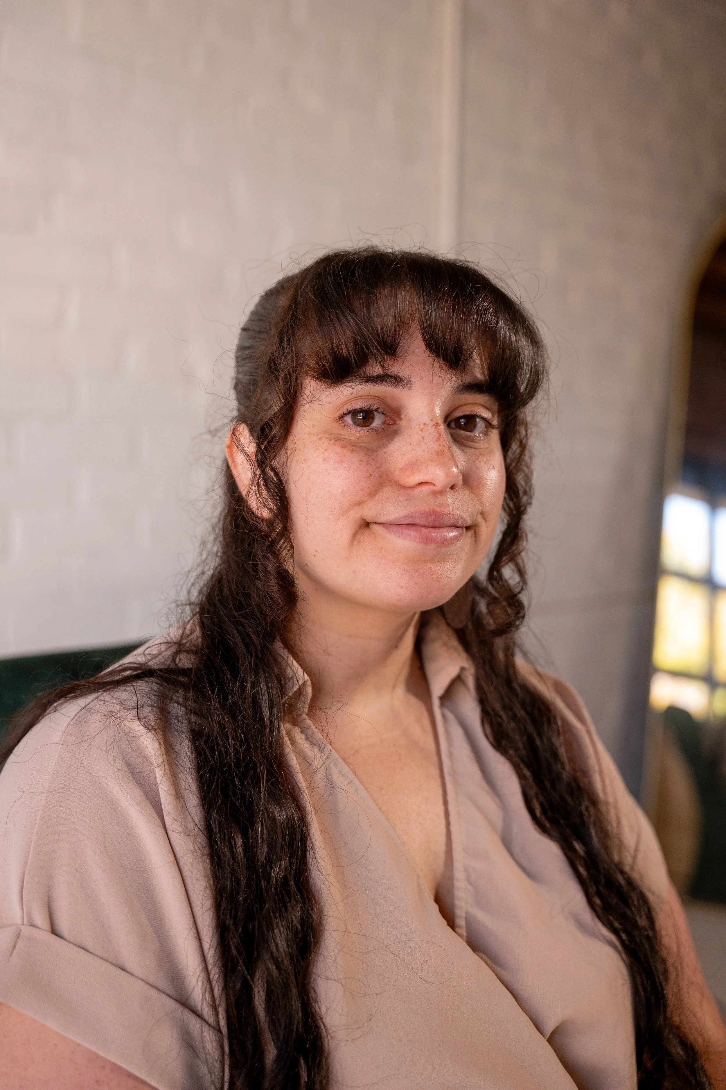 A woman with brown hair and freckles, wearing a beige shirt, sitting indoors near a window with blurred background.