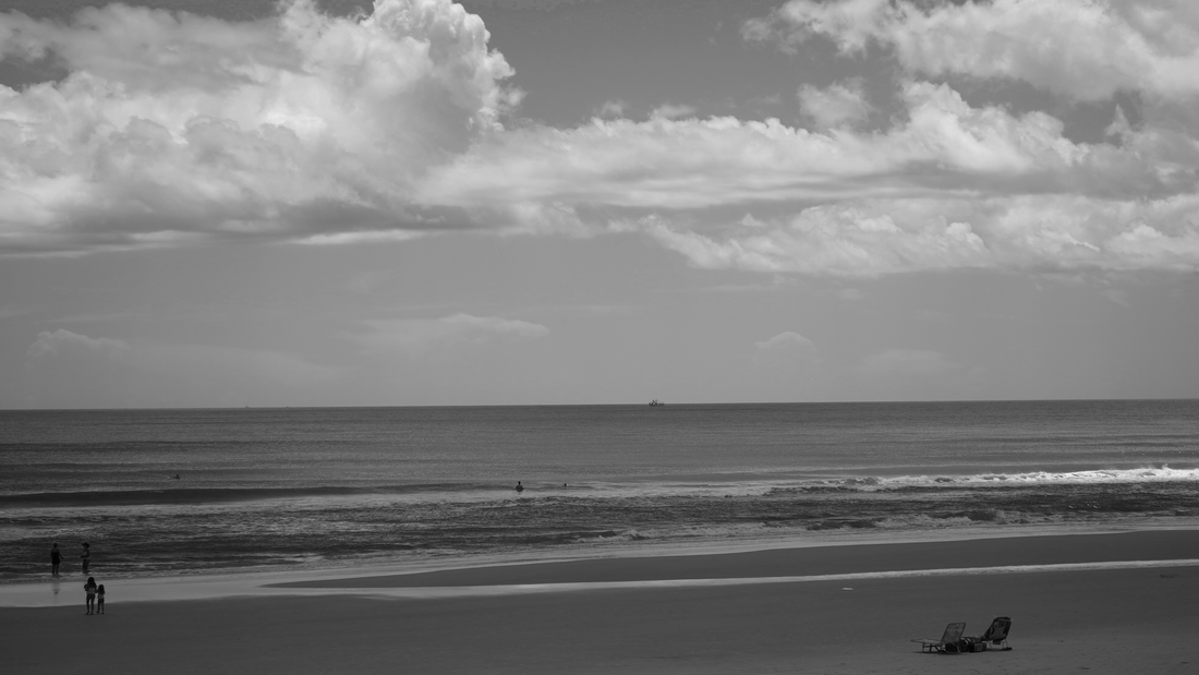 A black and white photo of a beach with a cloudy sky, calm ocean, and a few people walking along the shore. There are two lounge chairs positioned on the sand and a distant boat on the water.