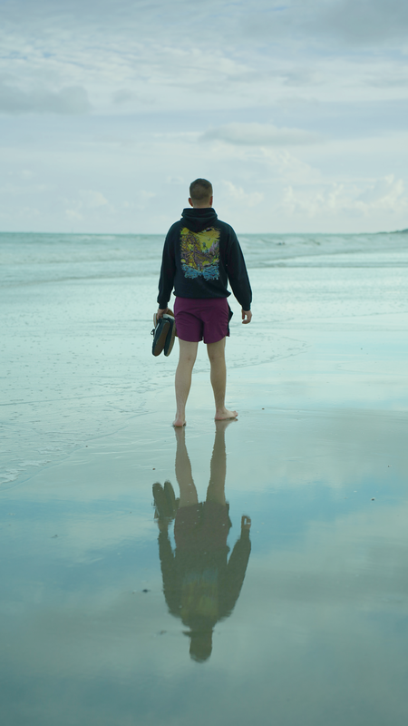 Person standing on wet beach holding shoes, facing the ocean during cloudy day with reflection in shallow water.