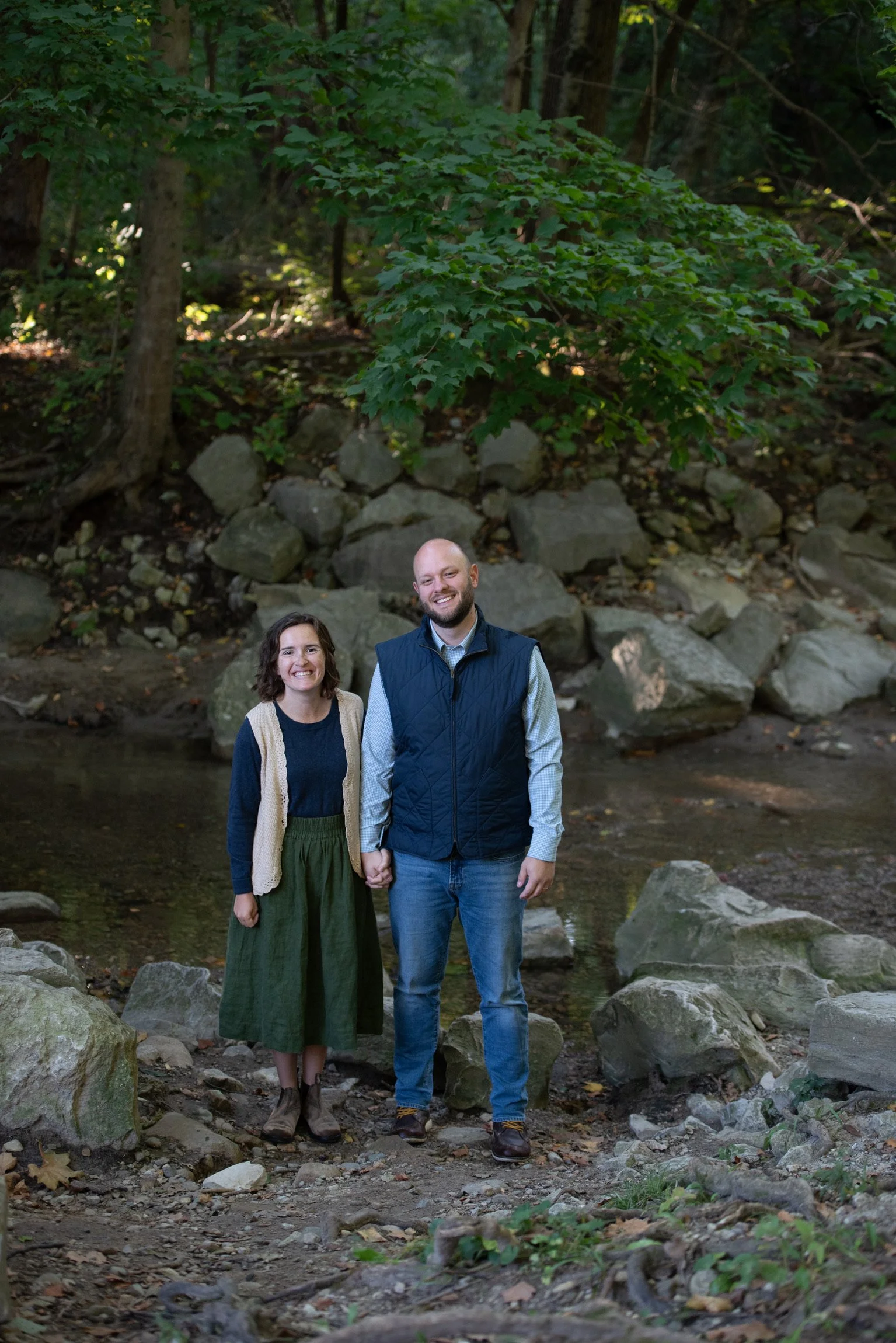 A smiling couple holding hands standing on rocks in a creek surrounded by trees and greenery in a forest.