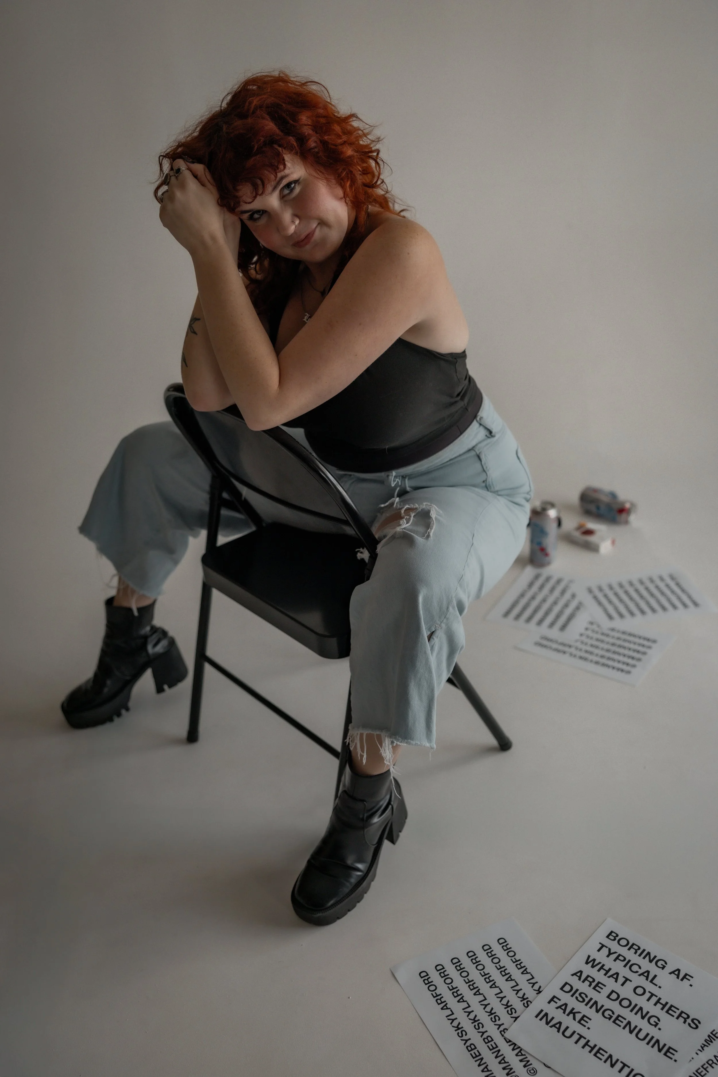 A woman with red curly hair seated on a black folding chair, wearing a black tank top, ripped light blue jeans, and black heeled boots, leaning forward slightly and resting her head on her hands, with a plain gray background and papers and soda cans on the floor behind her.