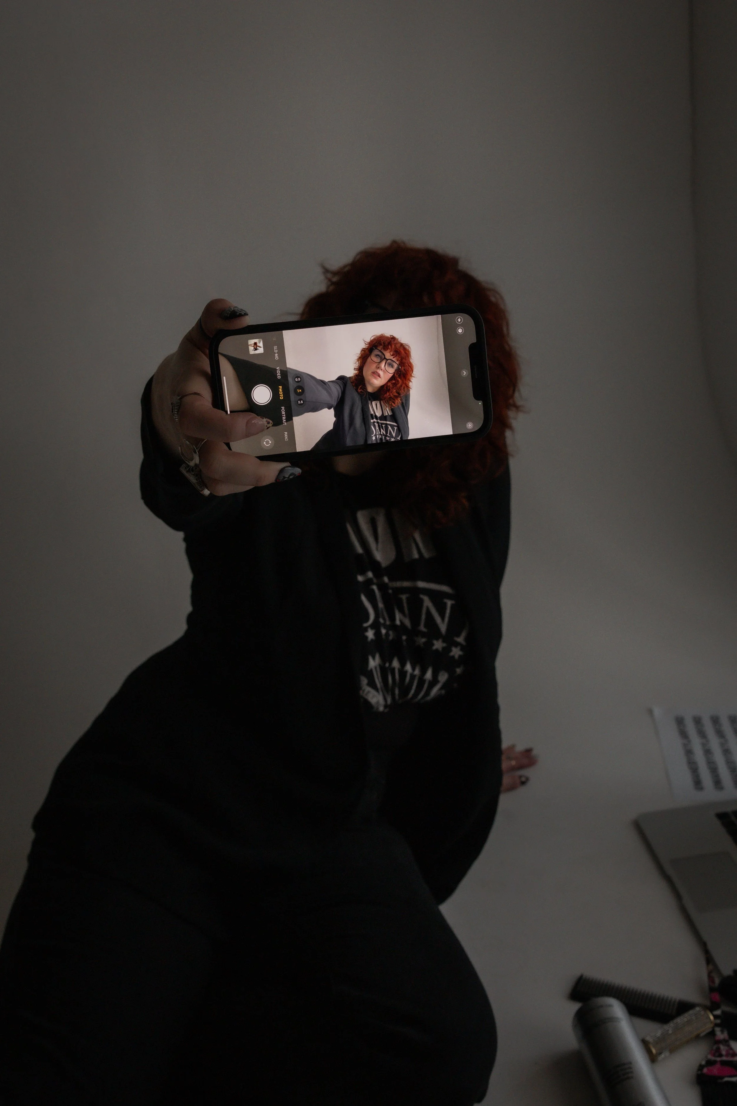 Person taking a selfie with a smartphone, with the screen showing their face and upper body, while the person is leaning back on a white surface, dressed in dark clothing with curly red hair and glasses.